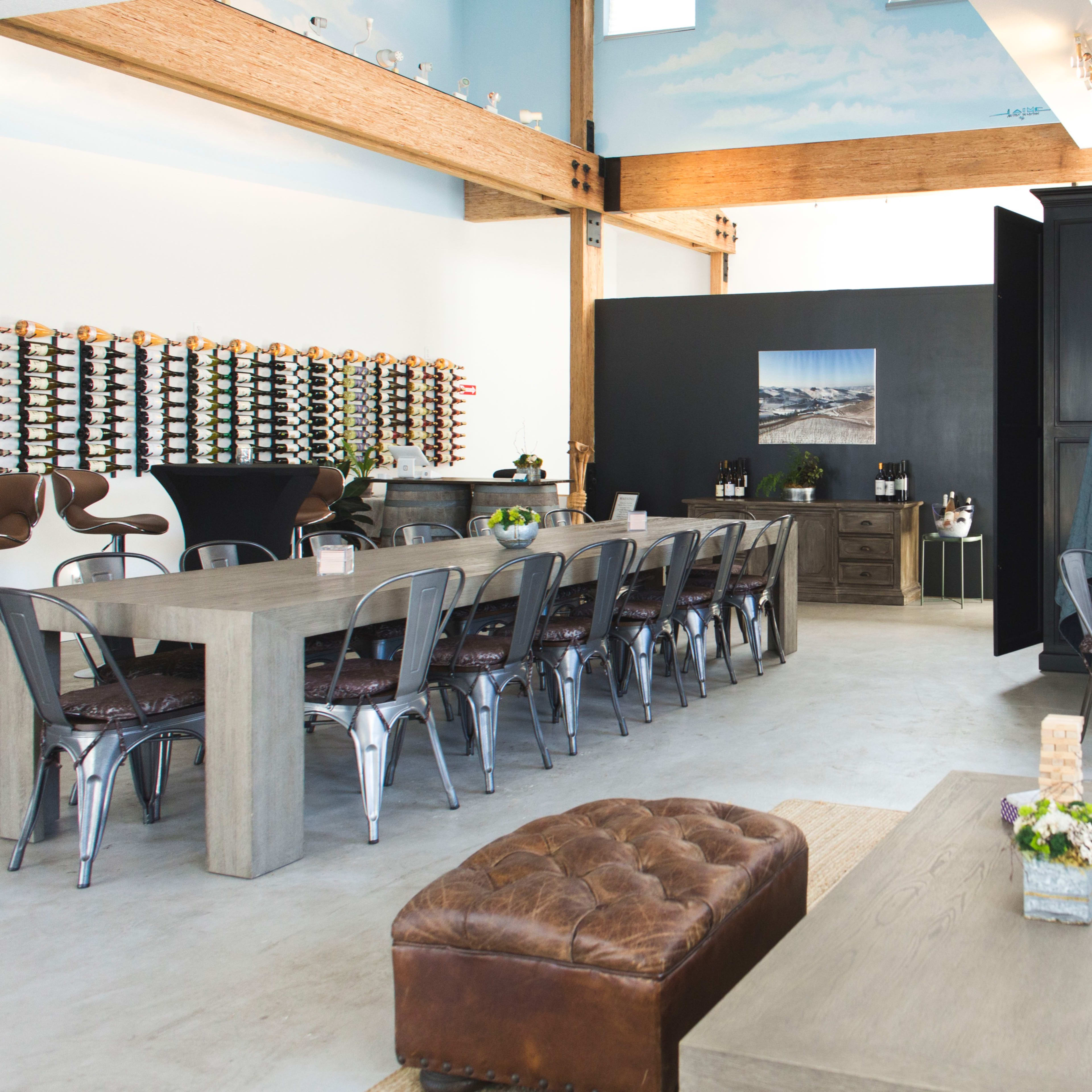 An indoor space featuring a long dining table, several chairs, and a bar area with bottles displayed on the wall.