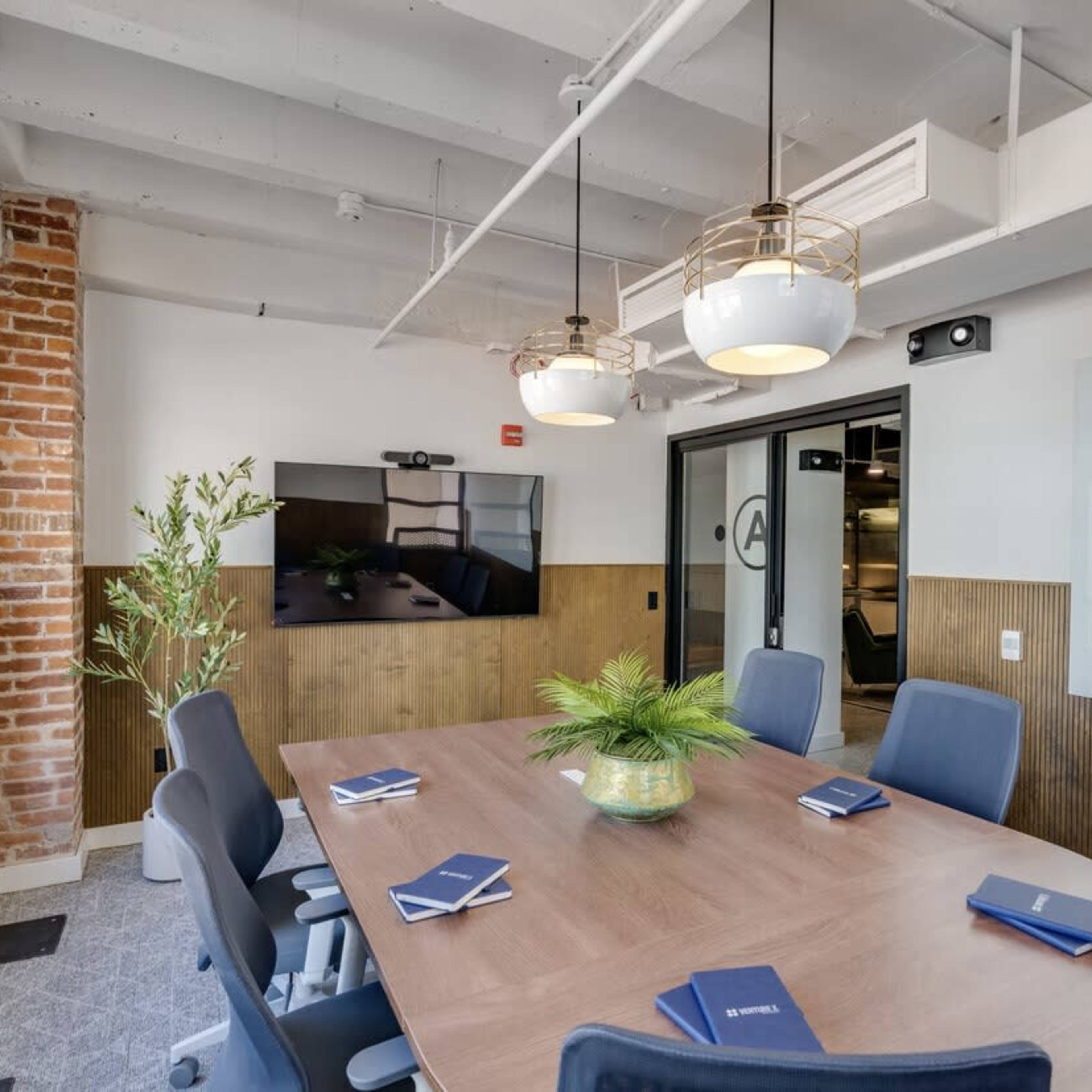 A modern conference room features a large oval table surrounded by blue chairs, with a television screen on one wall and decorative plants.