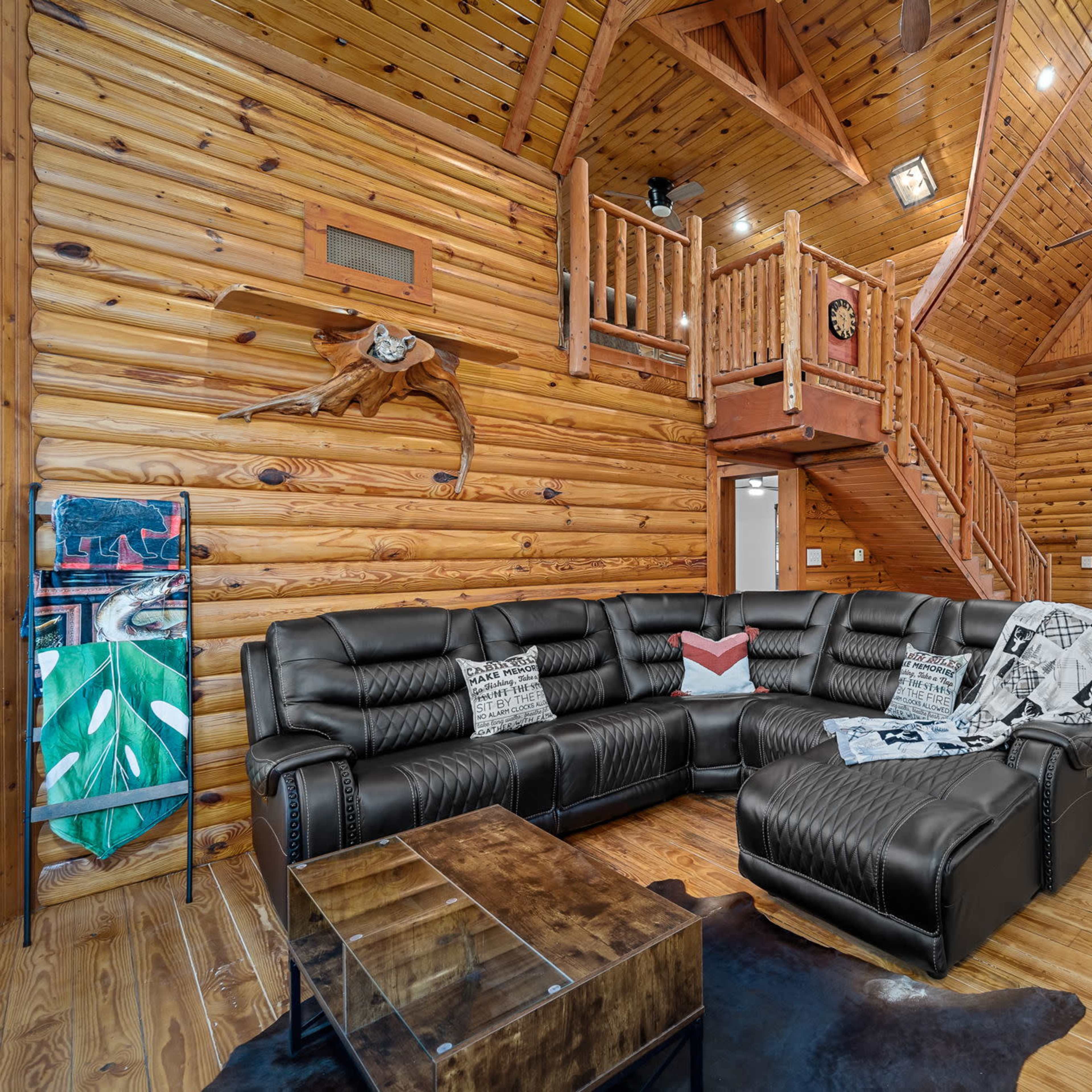A spacious wooden living room featuring a large black sectional sofa, a glass coffee table, and a wooden staircase leading to an upper loft area.