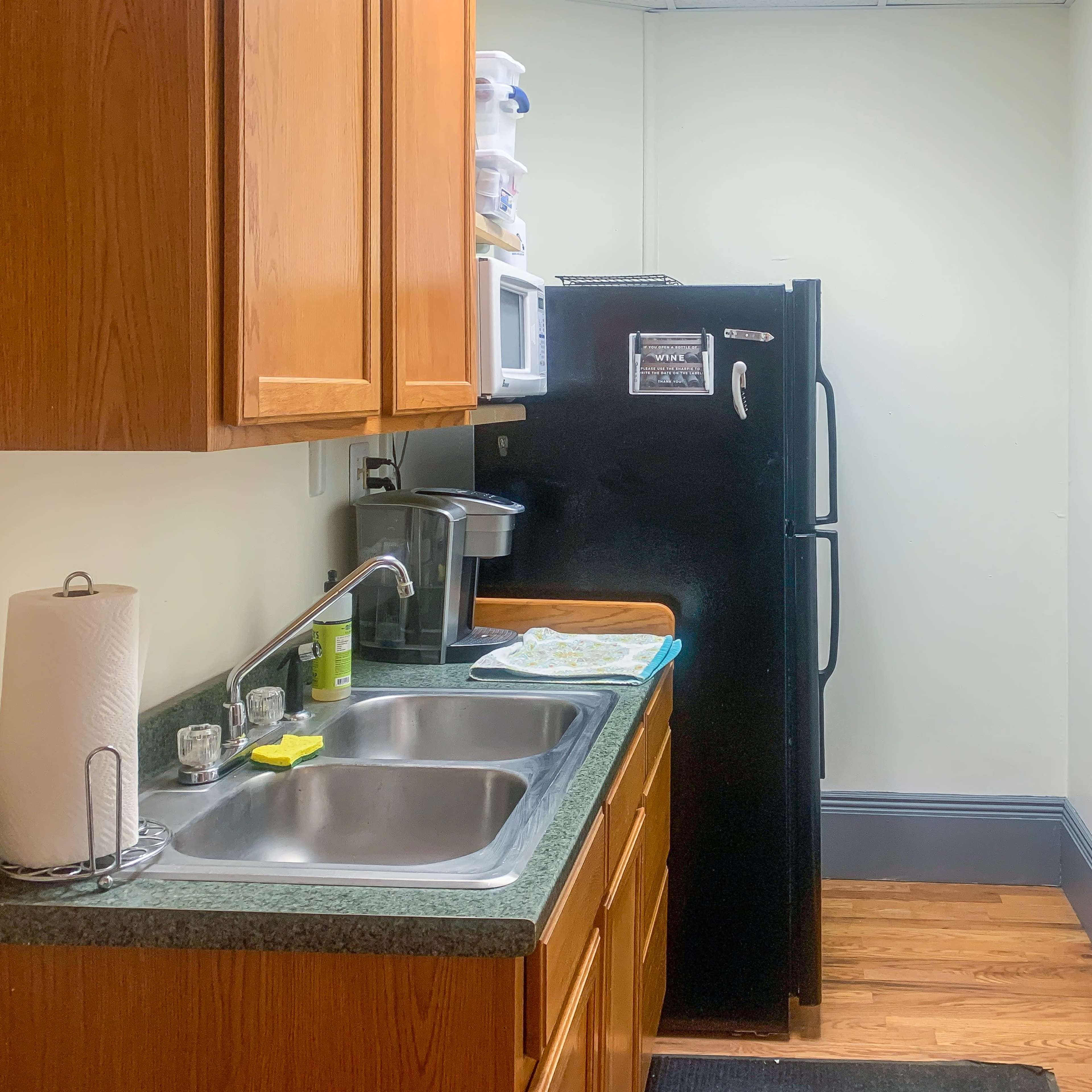 A small kitchen features wooden cabinets, a black refrigerator, a double sink, and a white shelving unit with various items.
