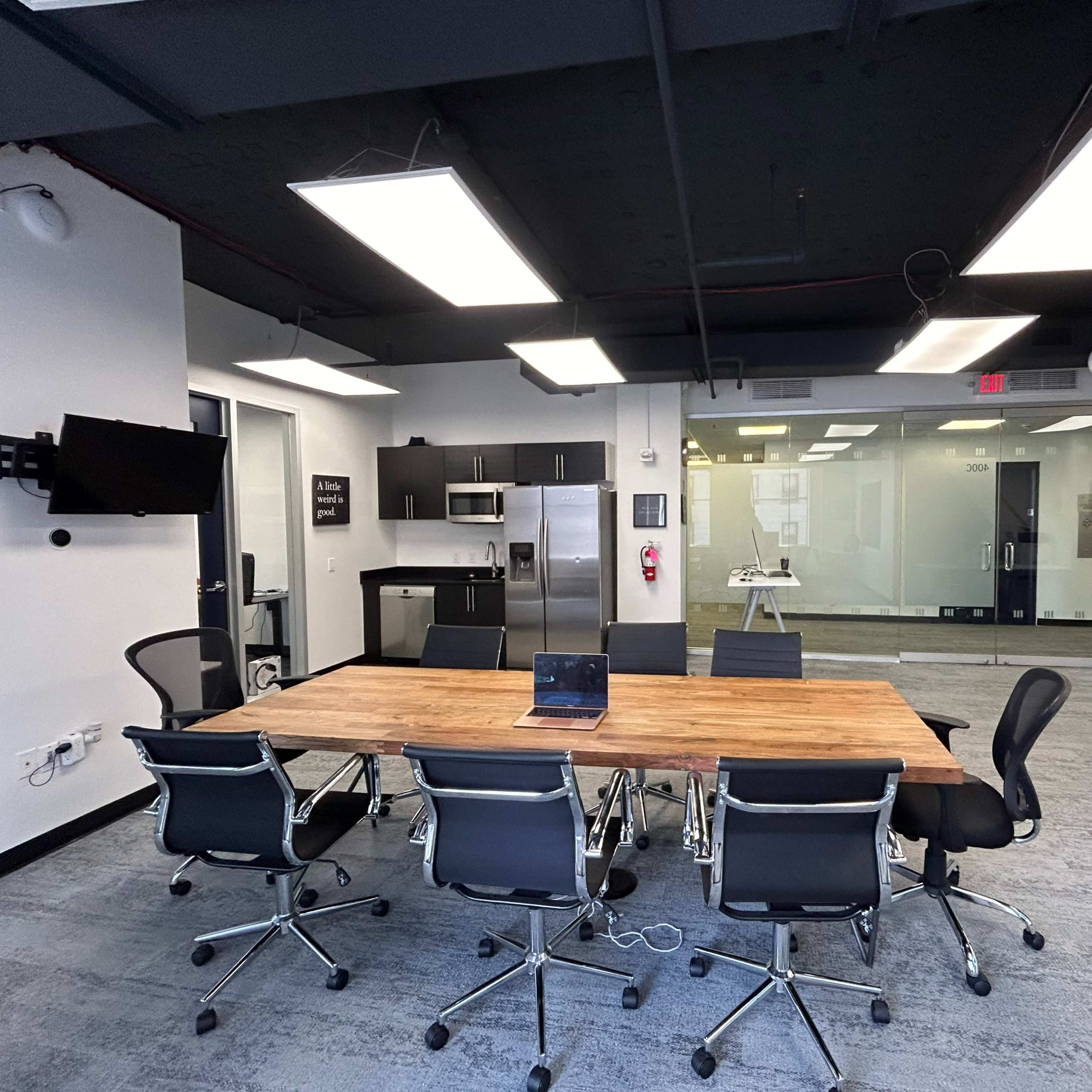A modern conference room with a large wooden table surrounded by black chairs, a kitchenette area, and glass walls.
