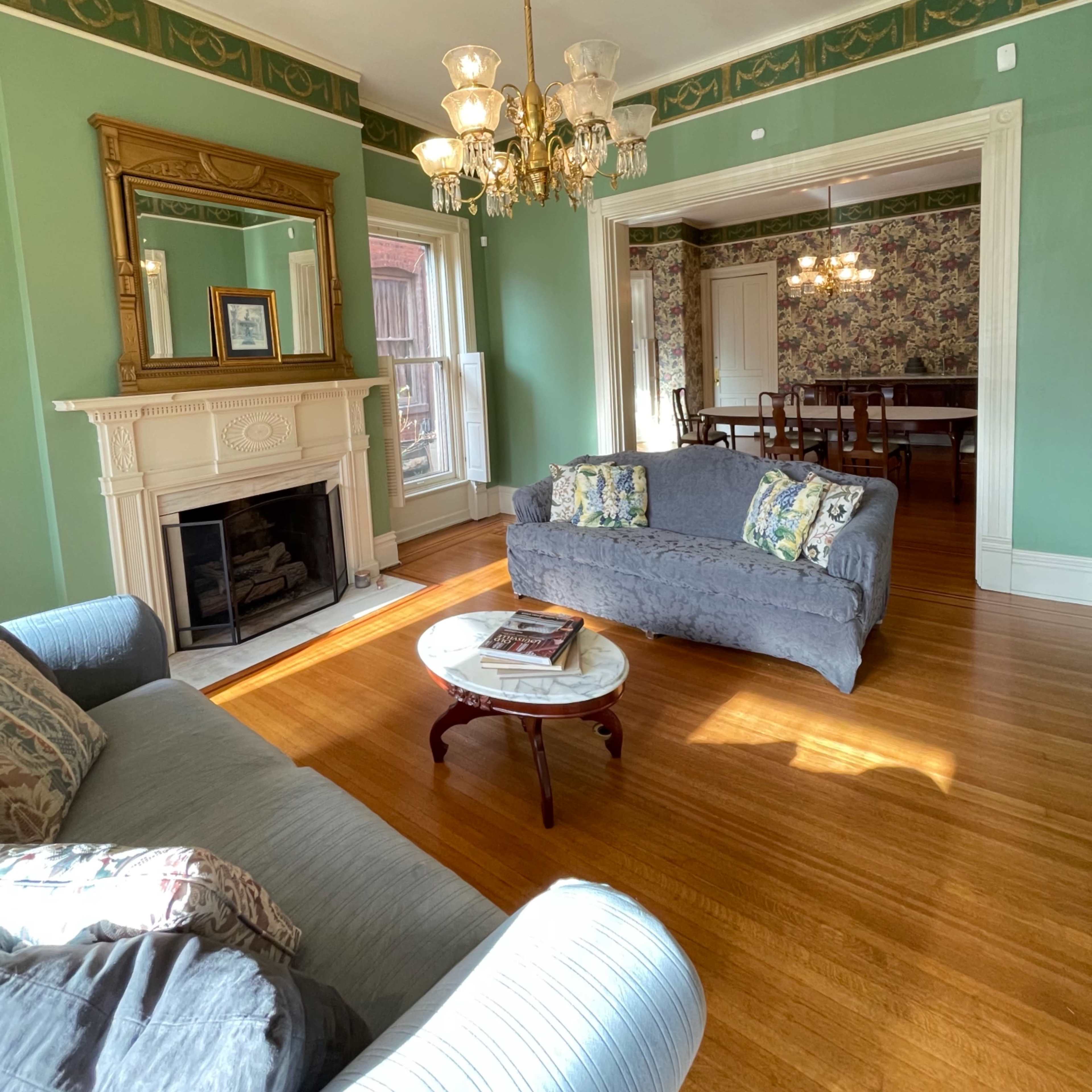 The room features a light green wall with a chandelier, a fireplace with a mirror above it, and a cozy seating area with two sofas and a round table.
