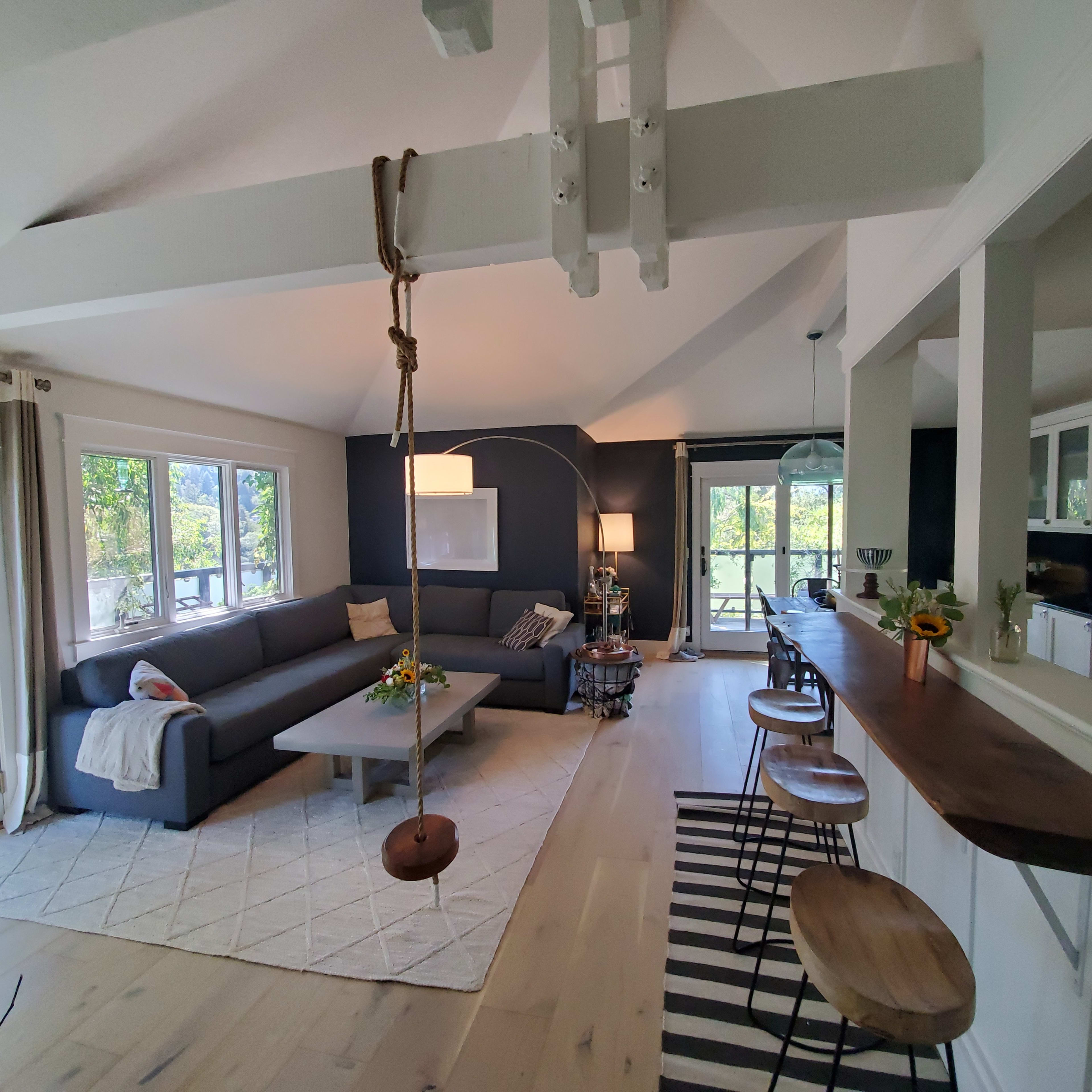 A modern living room features a gray sectional sofa, a wooden bar with stools, and a swing hanging from the ceiling, surrounded by large windows that provide natural light.