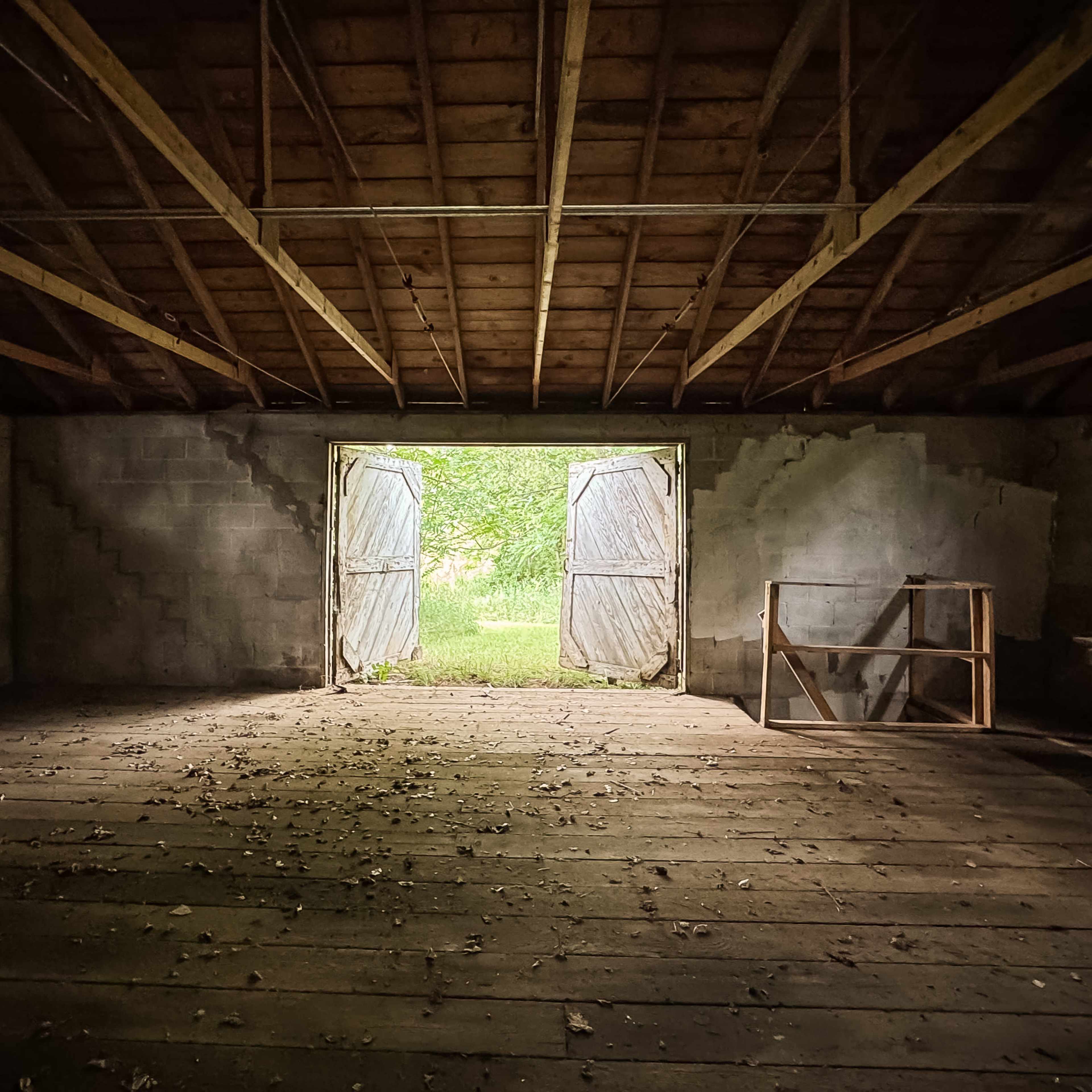 An empty barn interior with sunlight streaming through open double doors, illuminating the wooden floor covered in fallen leaves.