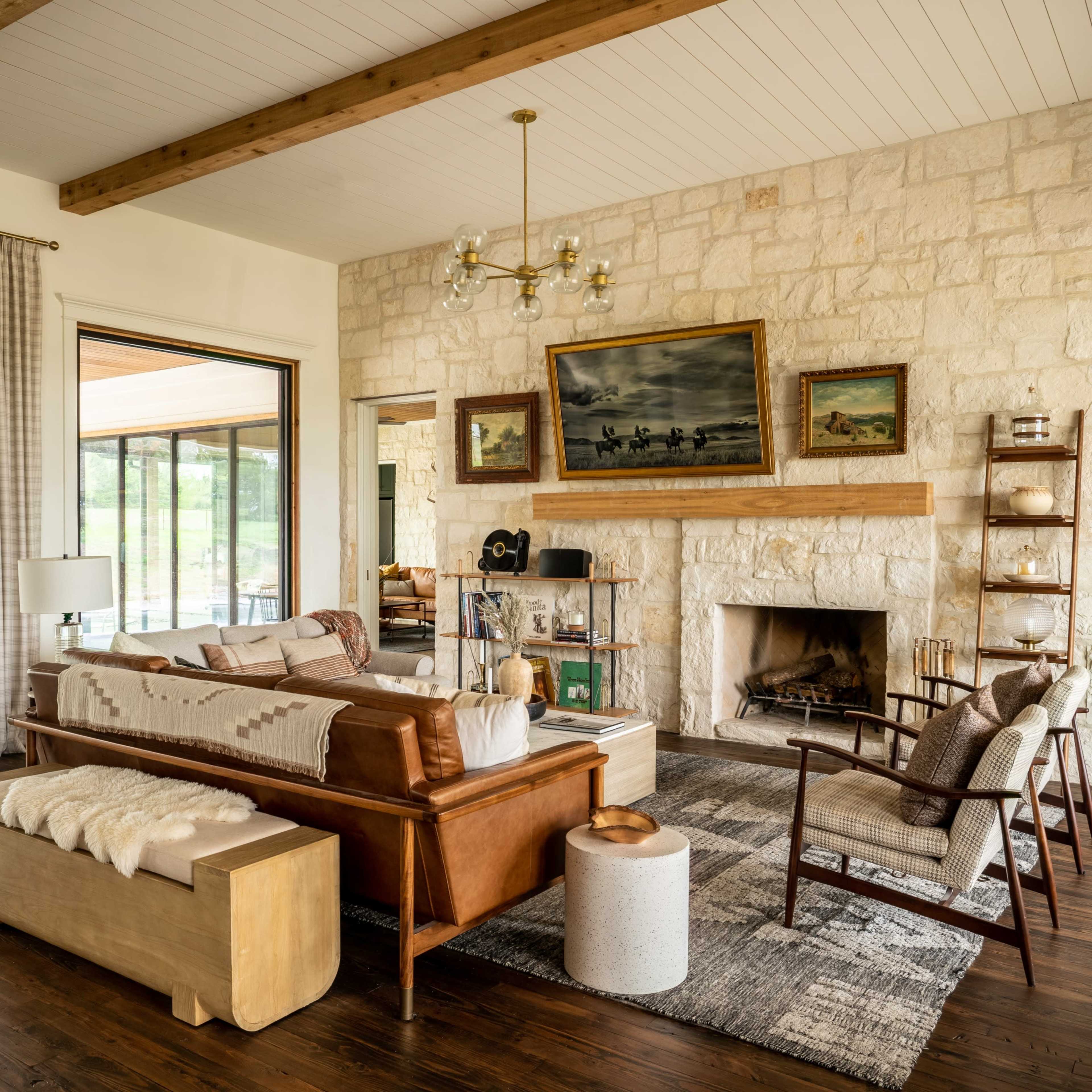 A modern living room features a stone fireplace, wooden beams, and a leather sofa, with large windows offering views of the outdoor patio.