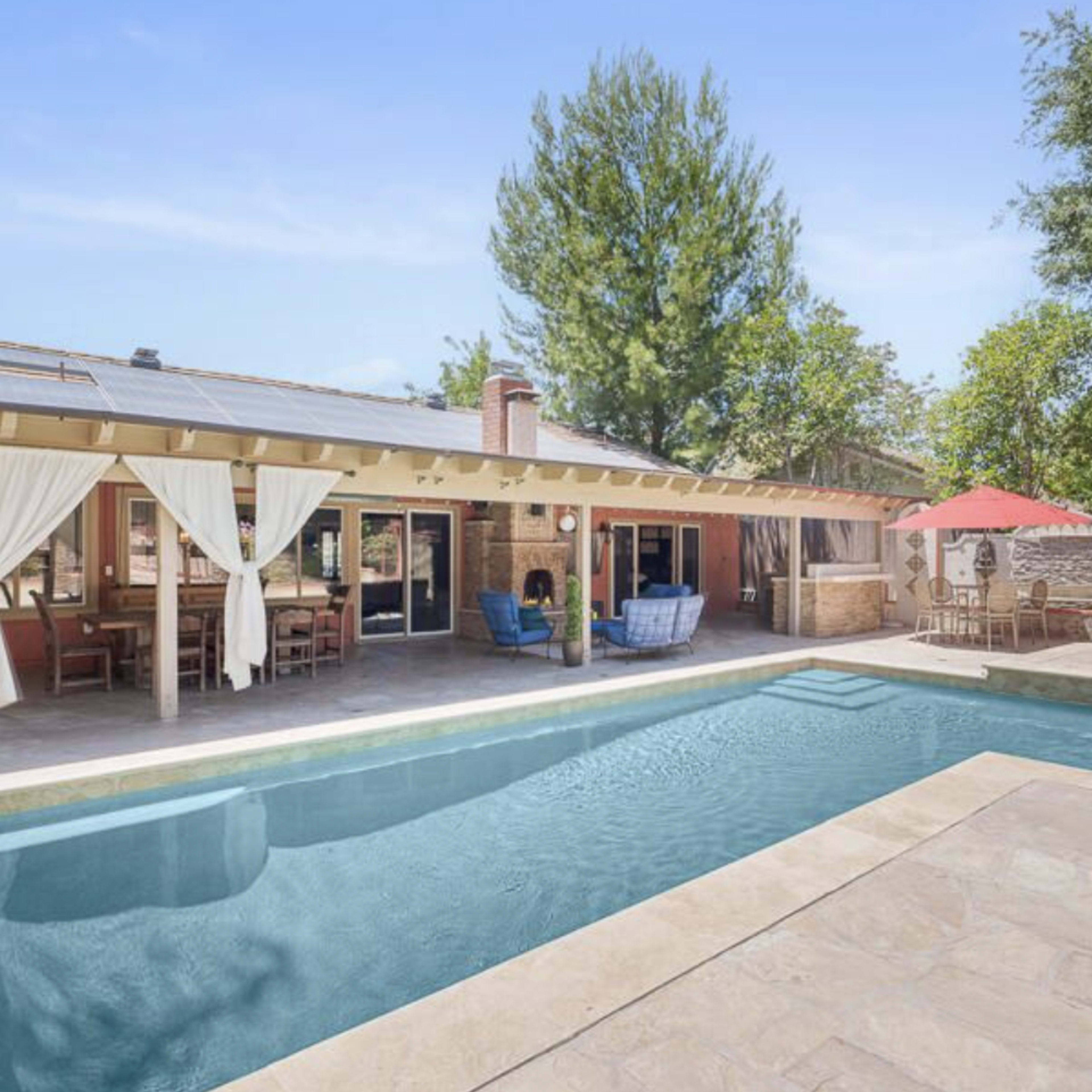 A backyard with a swimming pool, lounging area, and a covered patio with outdoor seating, surrounded by trees.