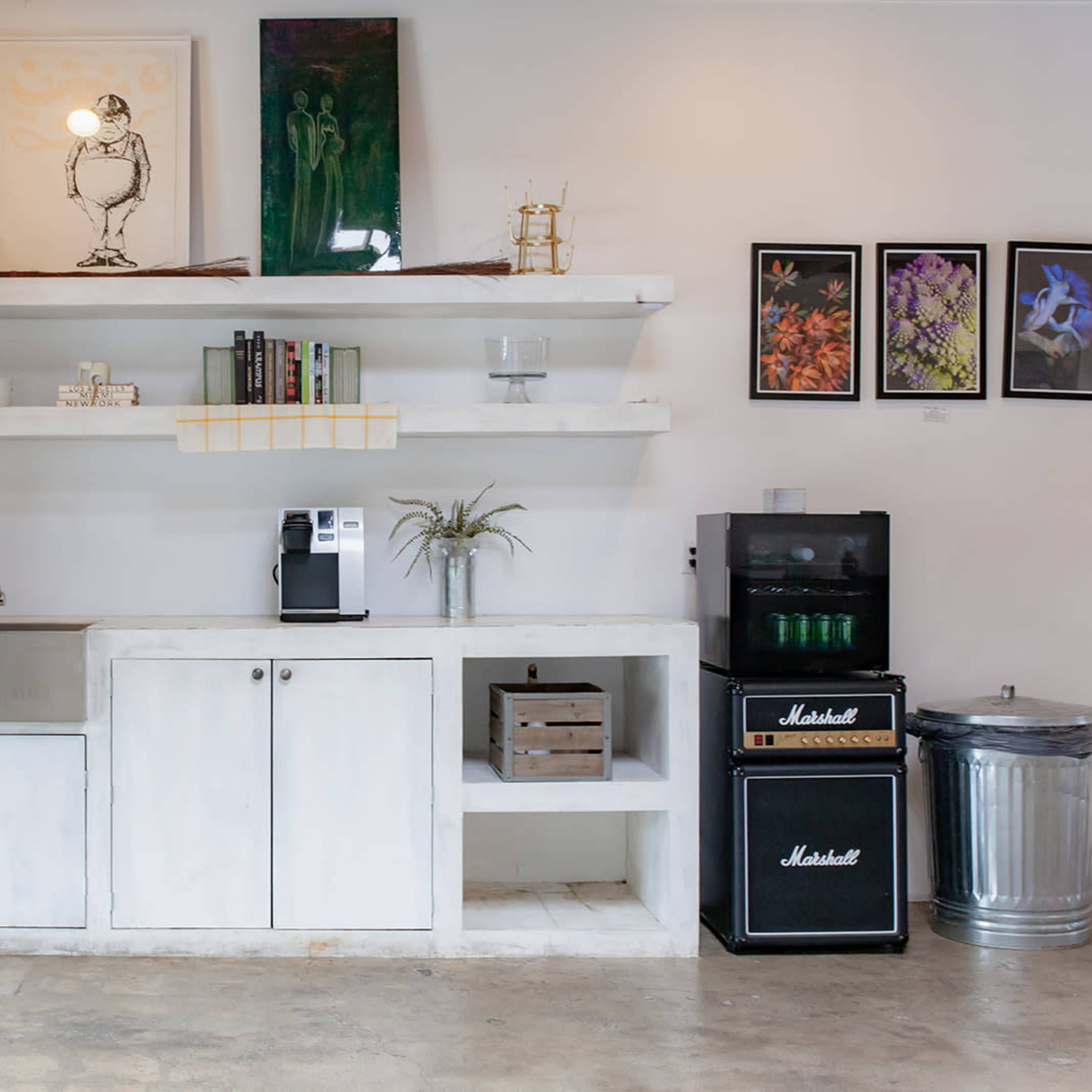 A minimalist kitchen area with white shelves, a stainless steel sink, a coffee machine, and various decorative items, set against a concrete floor.