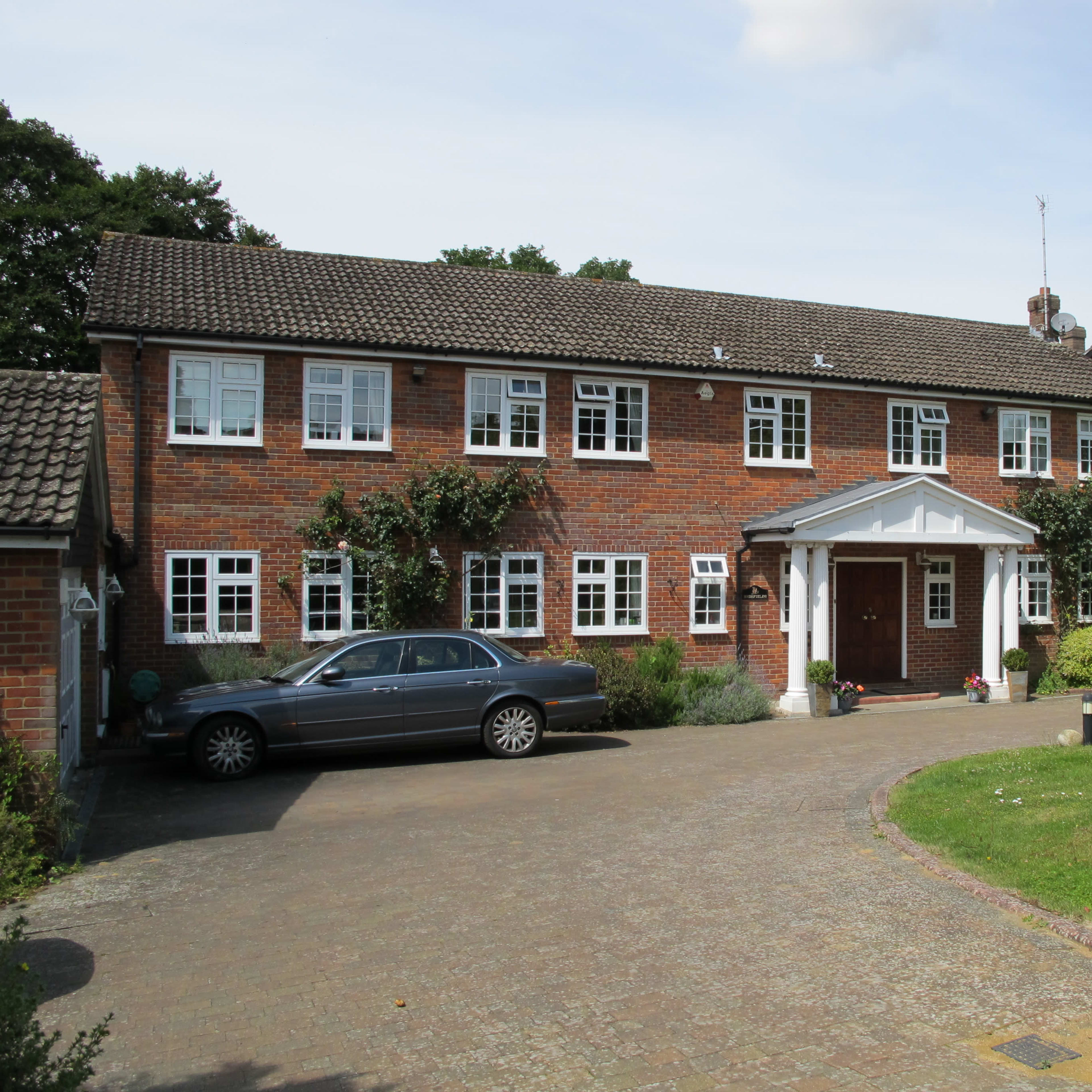 A brick house with white window frames and a covered entrance is set back from a stone driveway, alongside a parked silver car and well-kept landscaping.