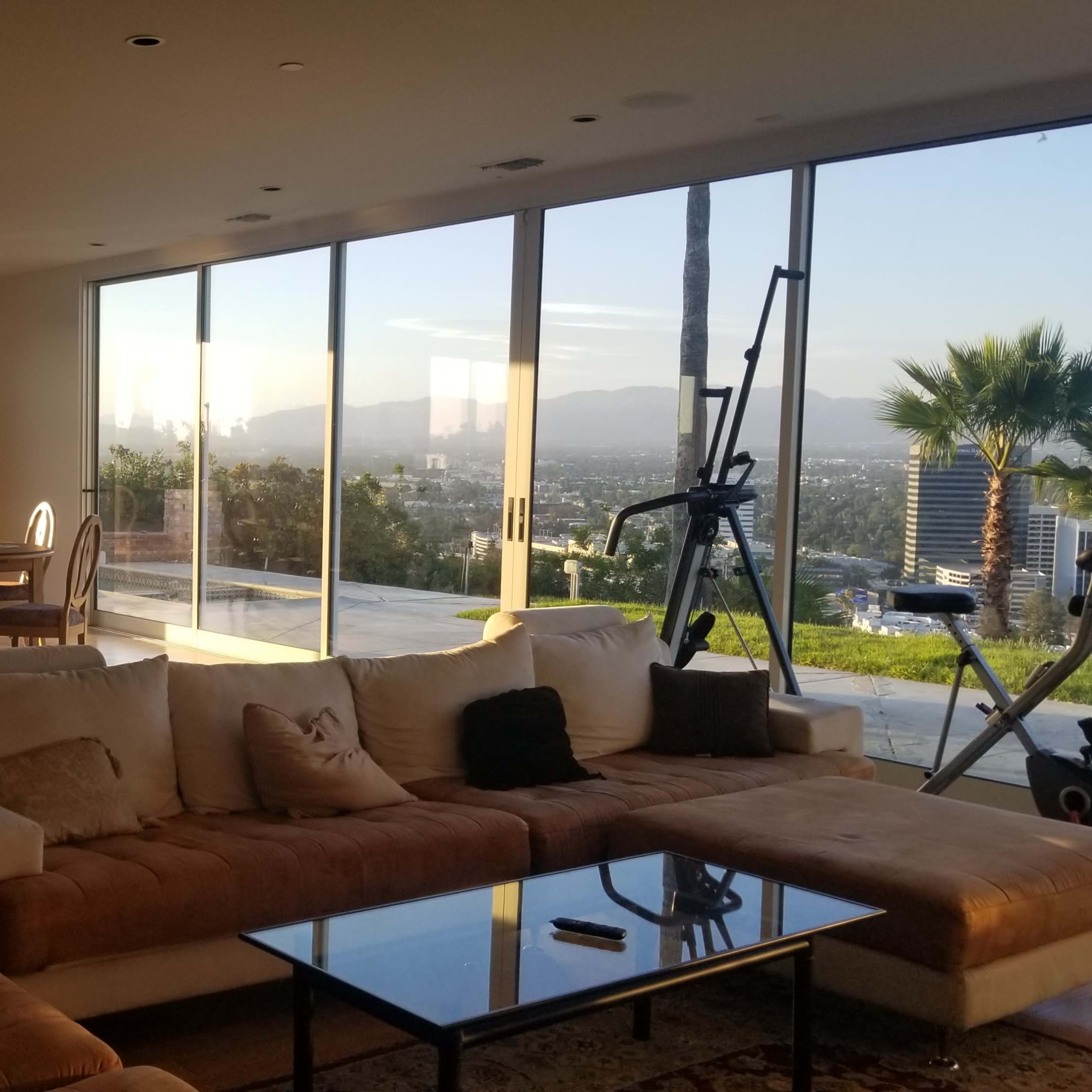 A modern living room with a large sectional sofa, a glass coffee table, and an open view of a balcony overlooking a cityscape at sunset.