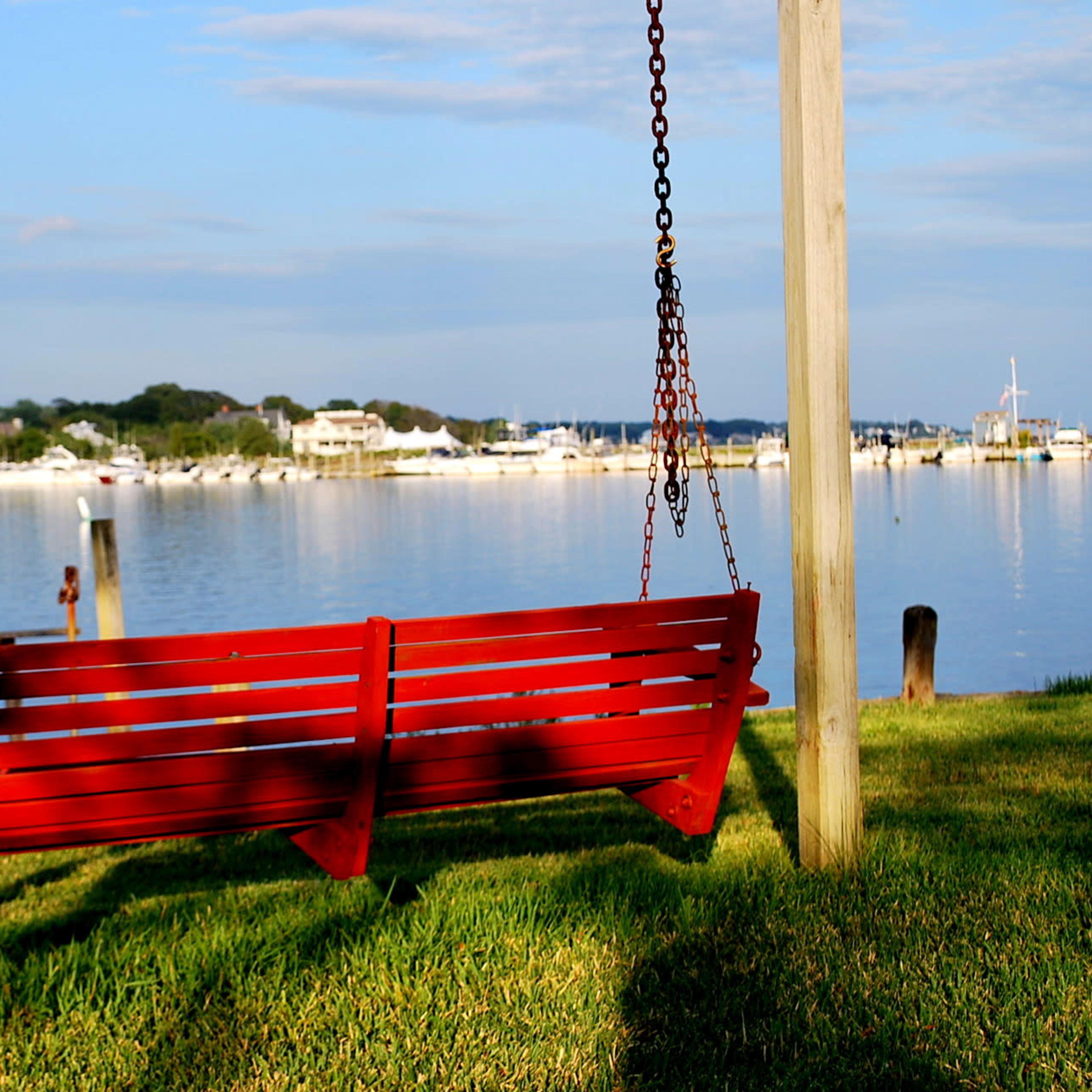 A red wooden swing hangs from a post overlooking a calm body of water with boats docked in the distance.