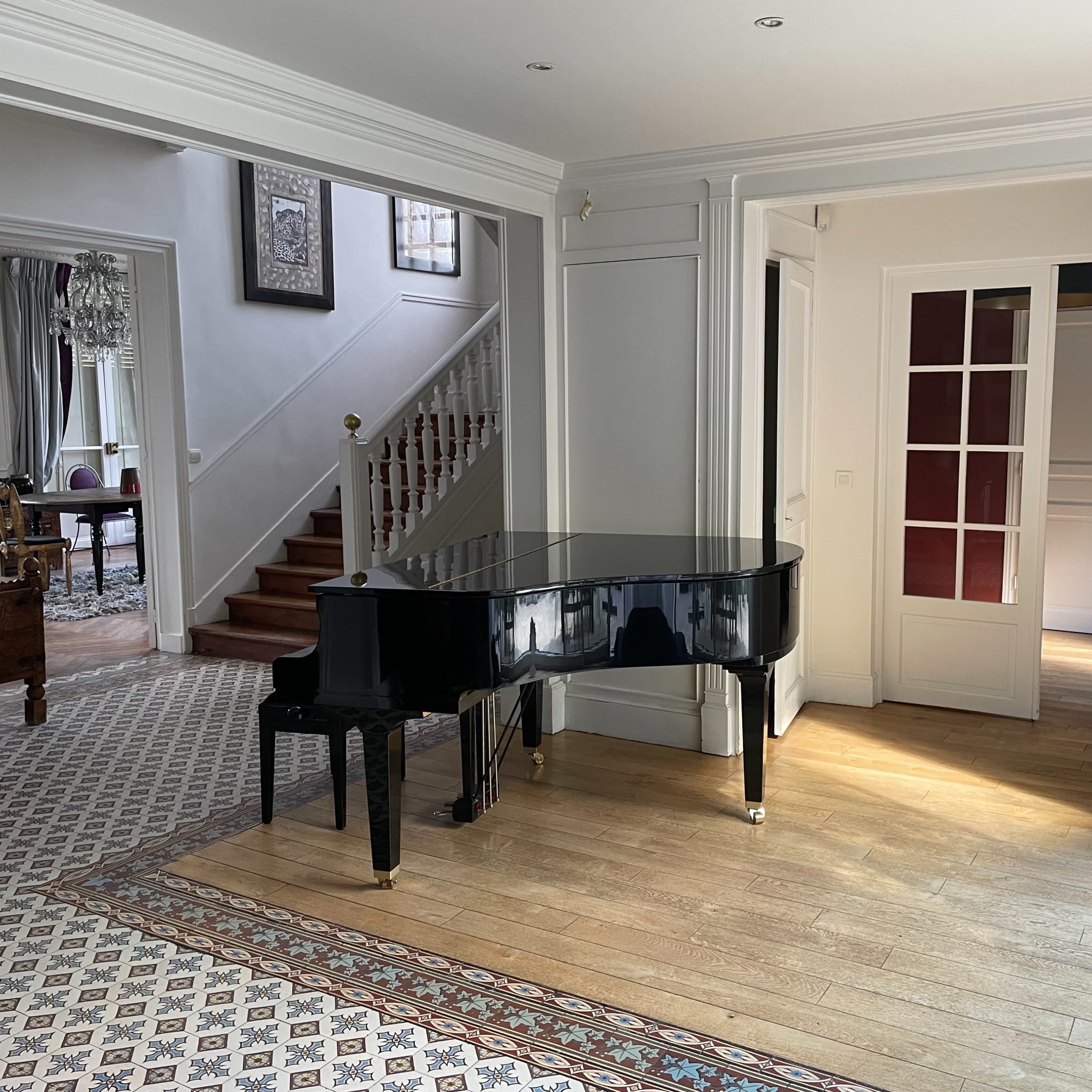 A spacious interior with a grand piano positioned centrally, surrounded by a patterned floor and stairway leading to an upper level.