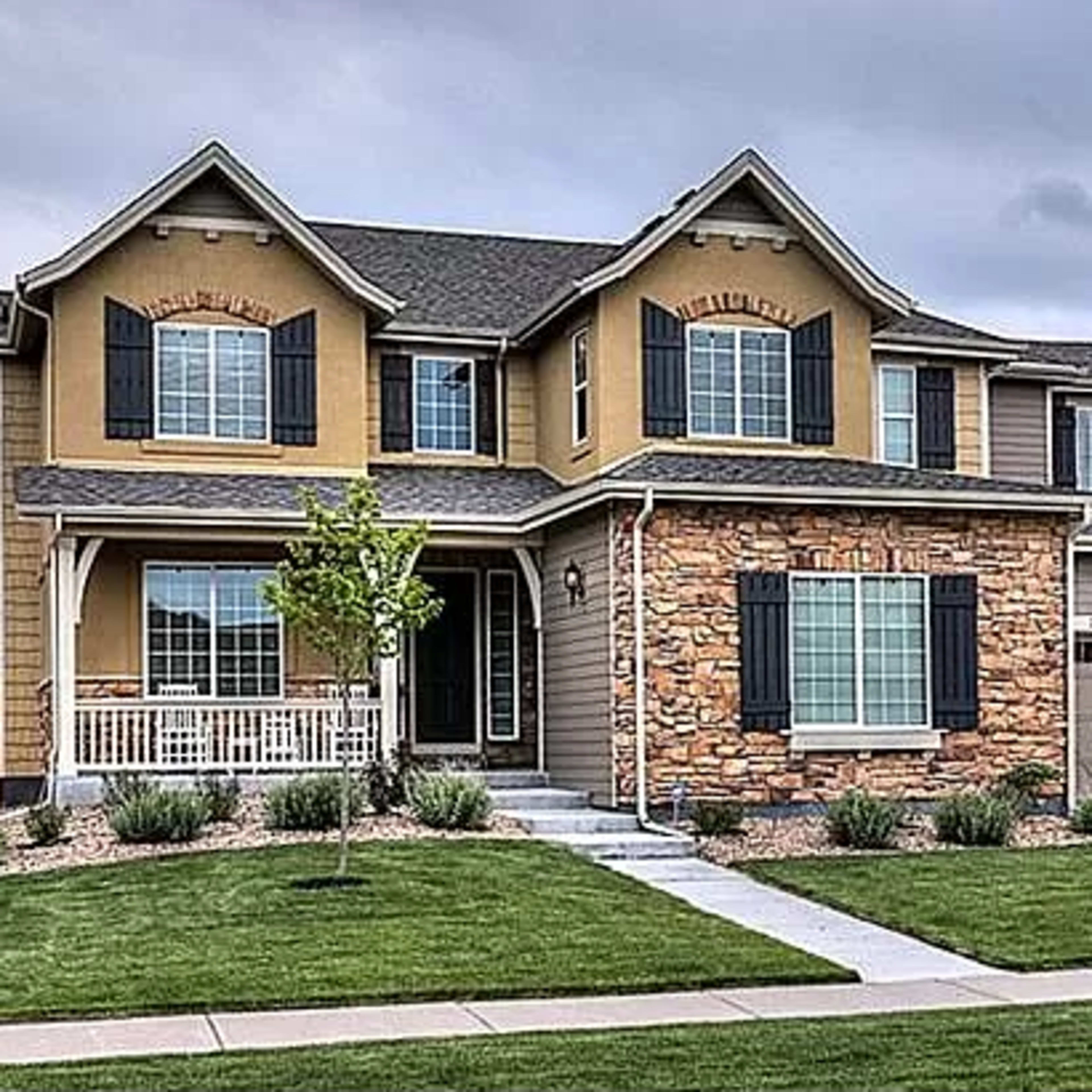 A two-story house features a mix of beige siding and stone with a front porch and a well-manicured lawn.