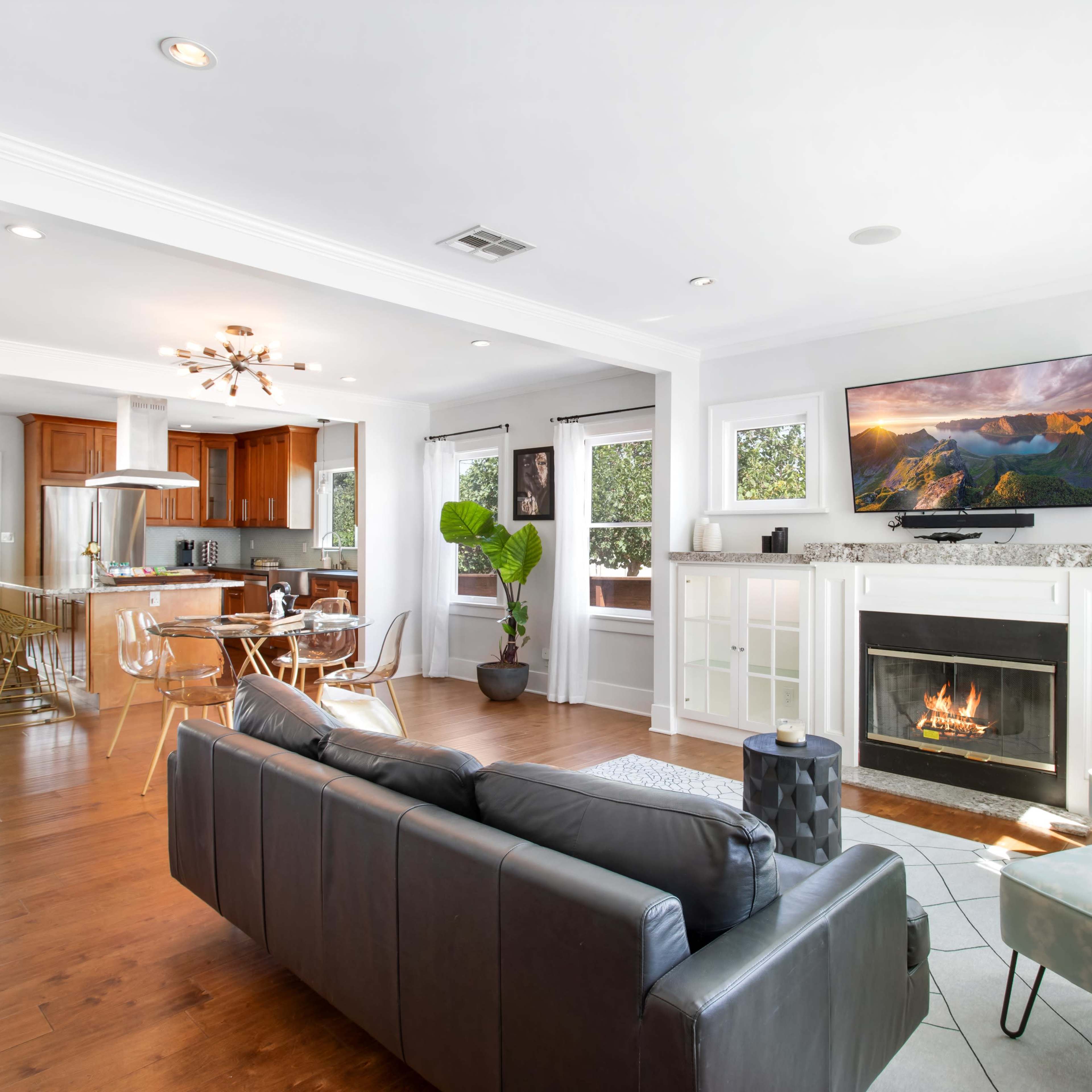A living room features a black sofa, a light blue armchair, a fireplace with a TV above it, and an open kitchen area with wooden cabinets in the background.