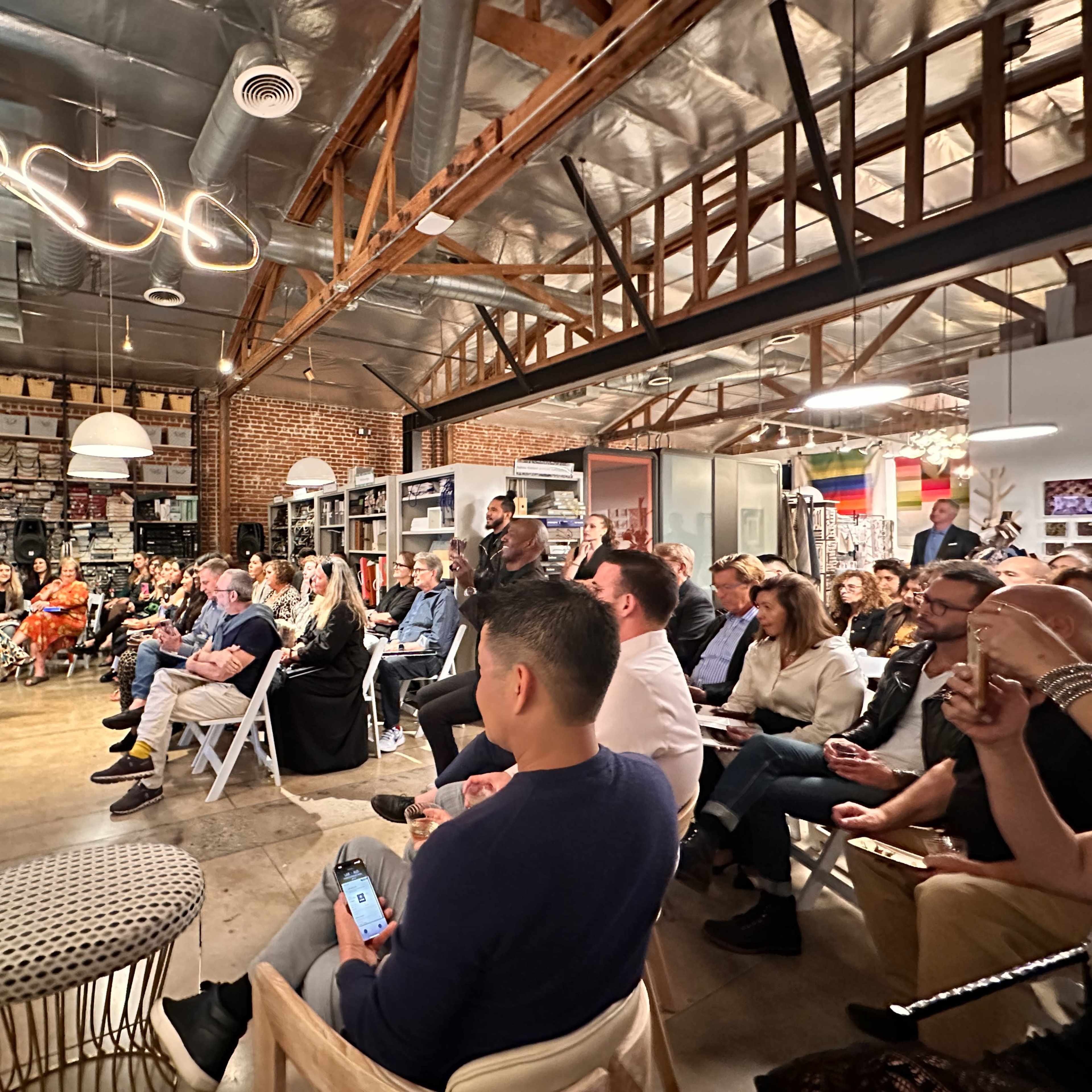 A diverse group of people is seated in a modern, industrial-style space, engaged in a discussion on a stage with a large screen displaying images.
