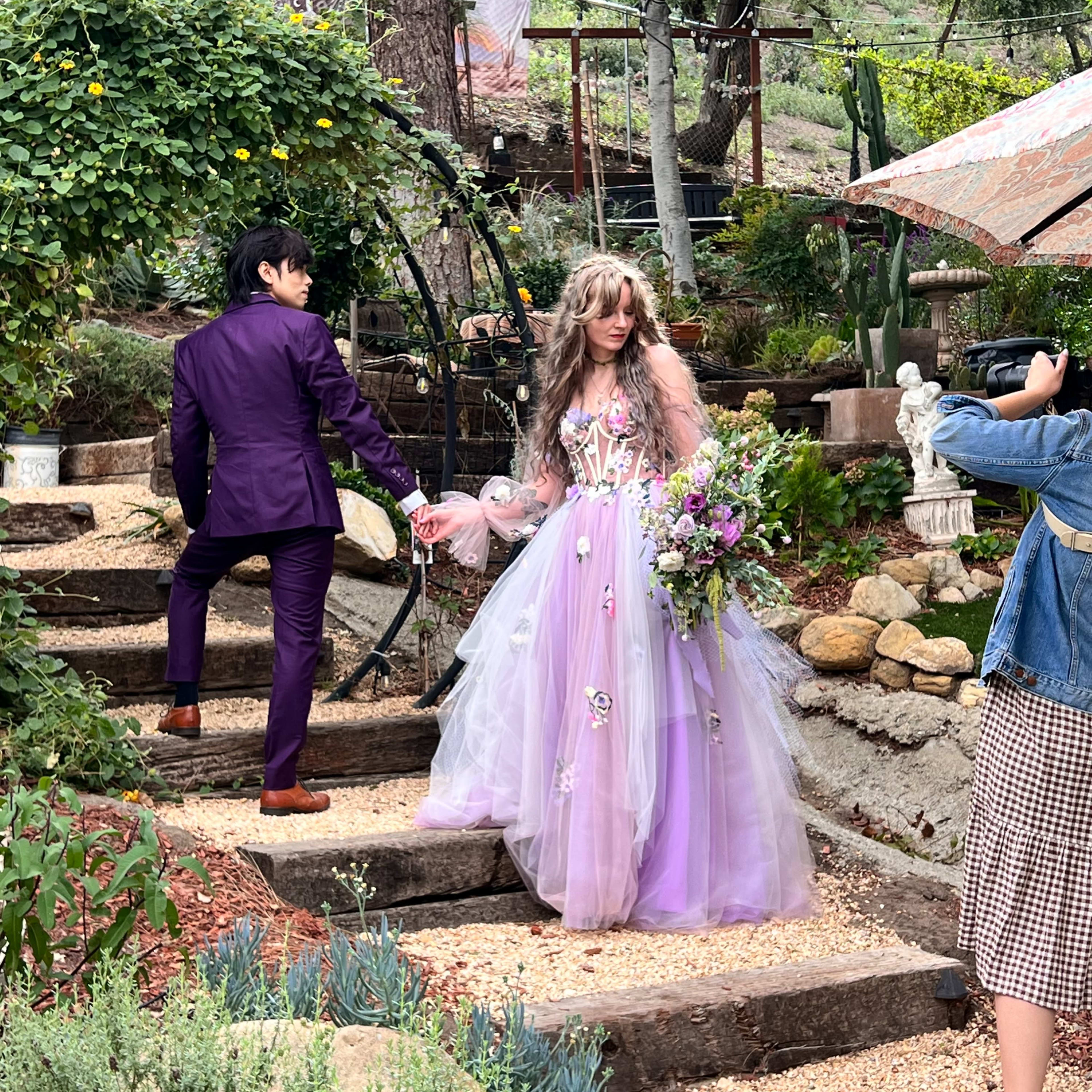 A couple dressed in formal attire poses on a pathway surrounded by greenery and decorative elements, while a person takes a photo of them.