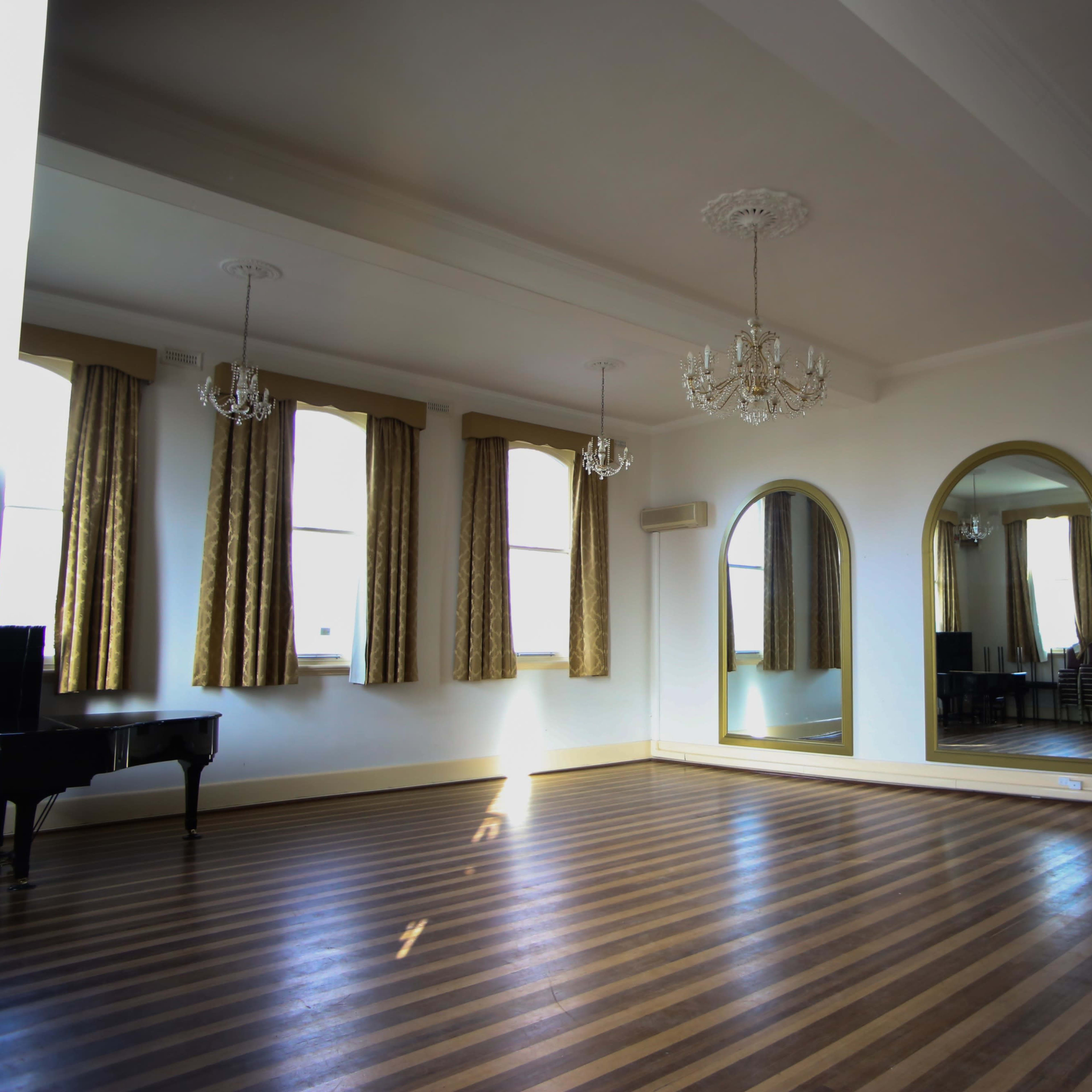 A spacious dance studio with wooden striped flooring, large mirrors, and elegant chandeliers.