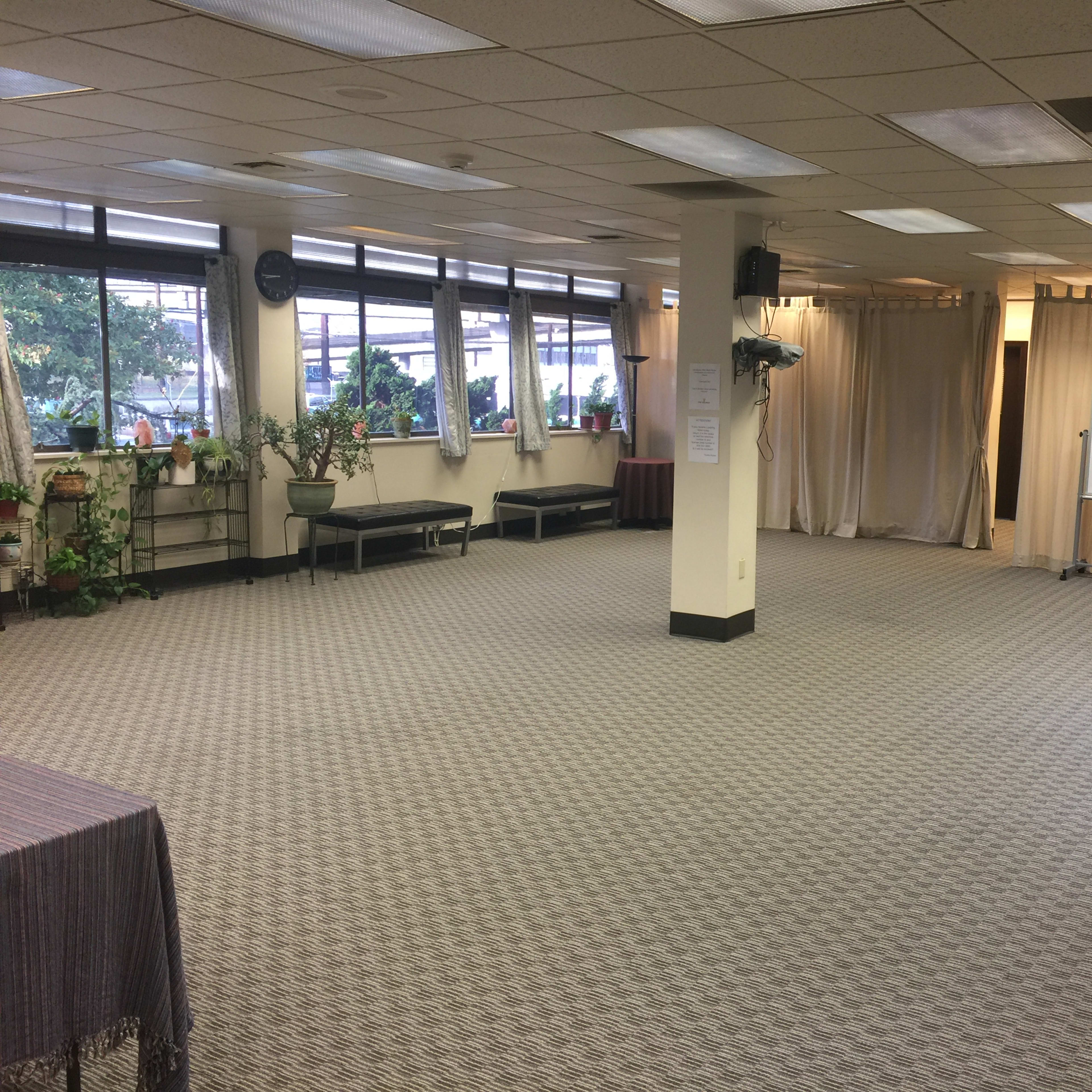 A large, empty room with windows on one side, plants along the walls, and a whiteboard positioned against a curtained backdrop.