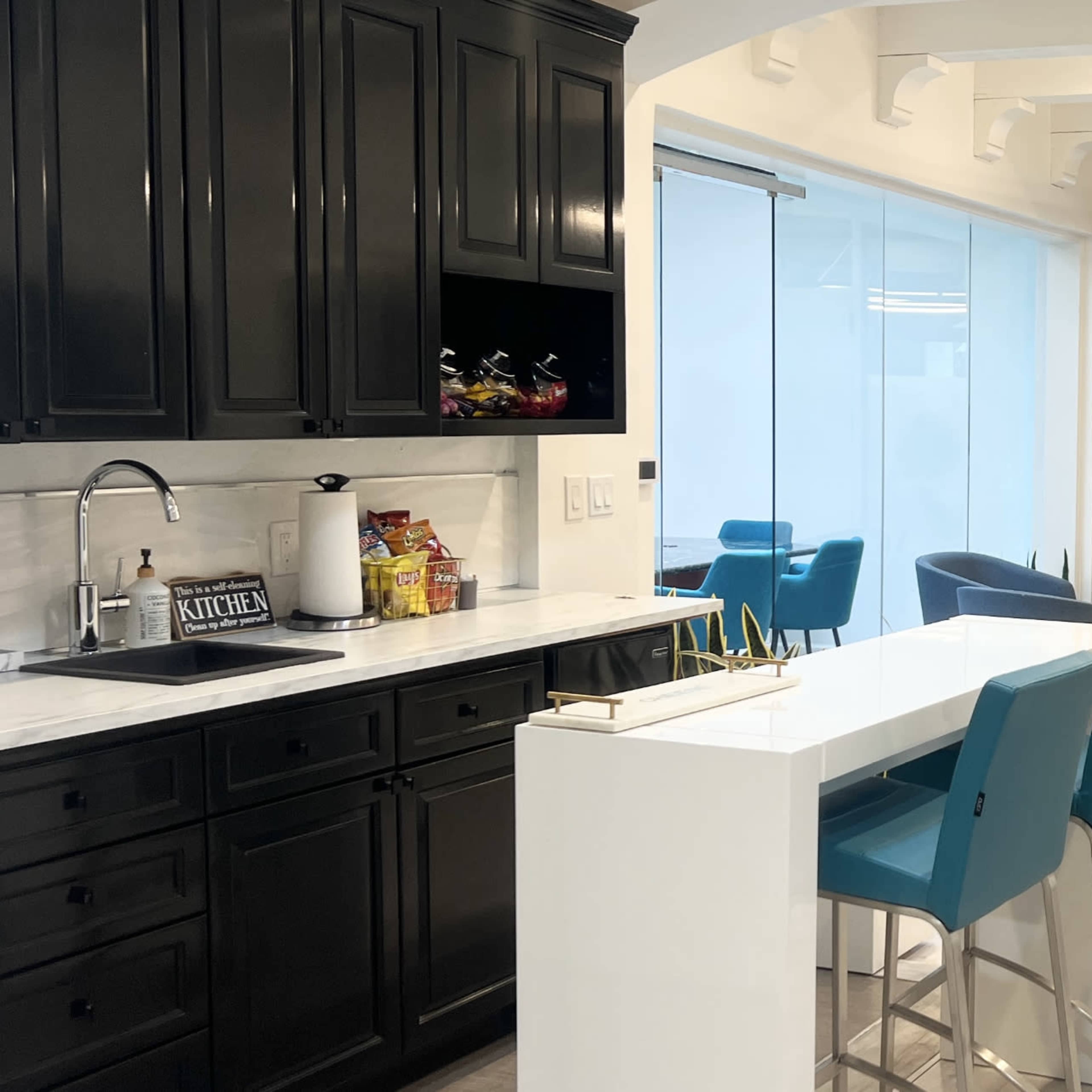 A modern office interior featuring a kitchenette with dark cabinetry, a white island with blue stools, and a lounge area with blue chairs and potted plants.