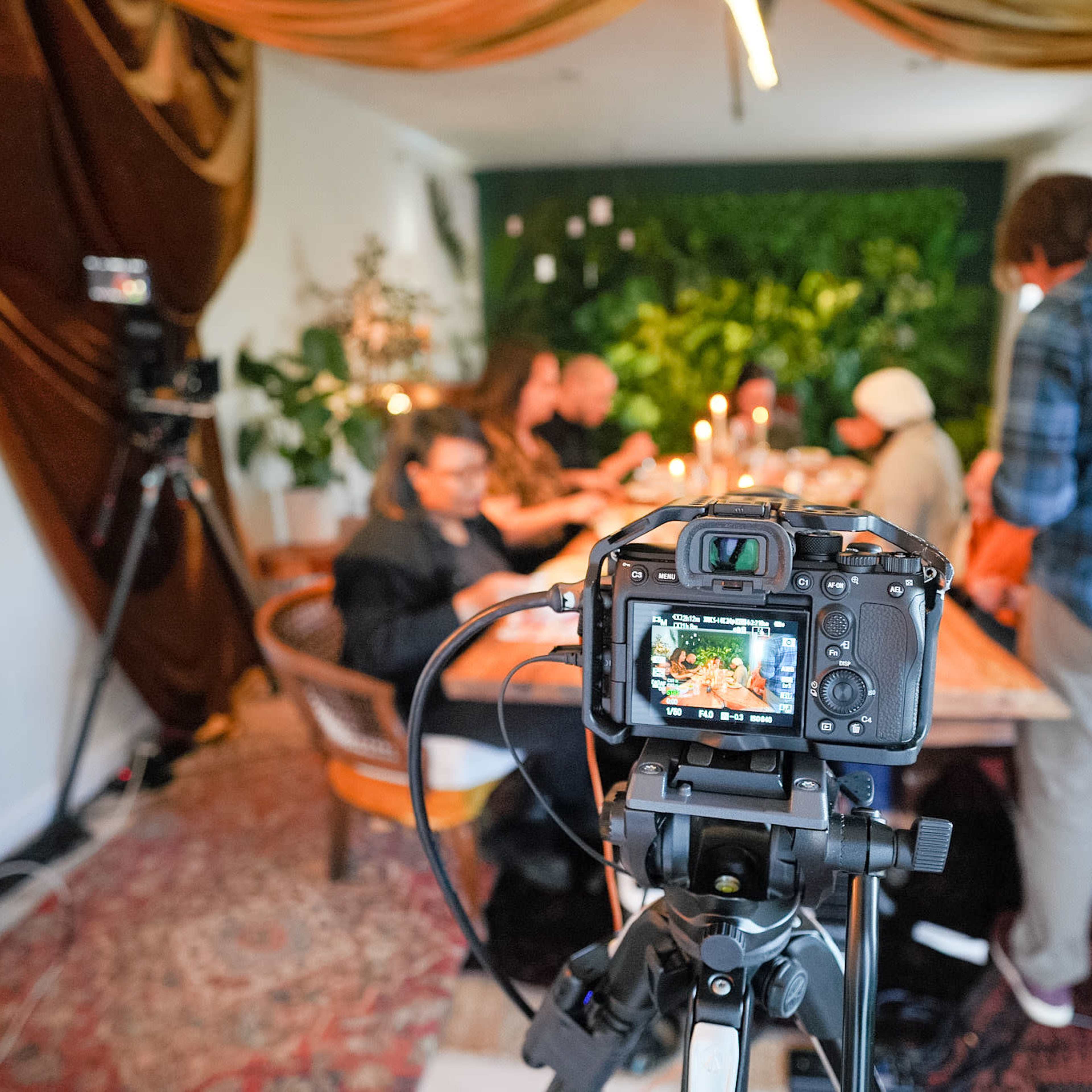A camera on a tripod focuses on a dining table surrounded by people, with greenery and decorative drapes in the background.