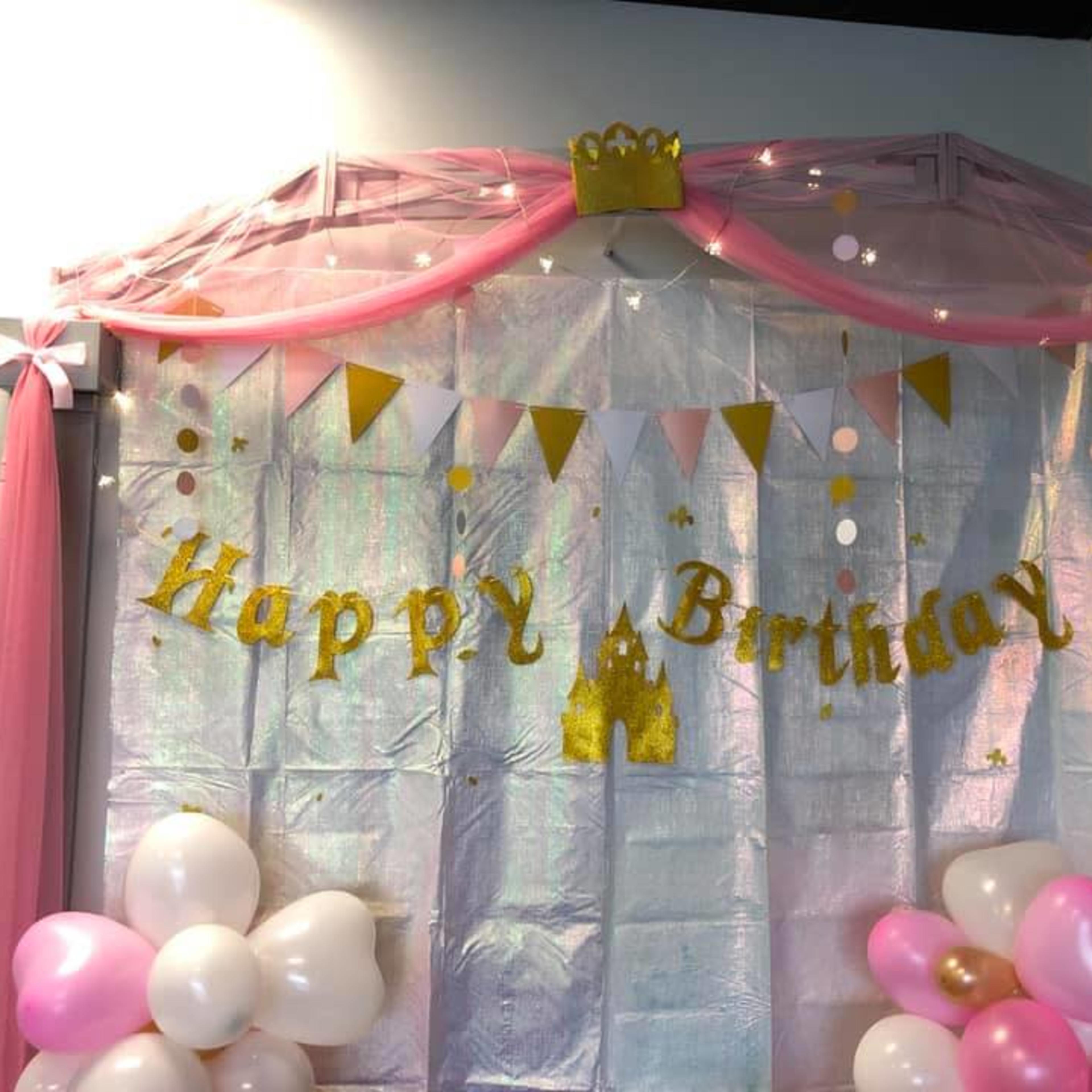 A colorful birthday party backdrop with a "Happy Birthday" banner, pink and gold decorations, and balloons.