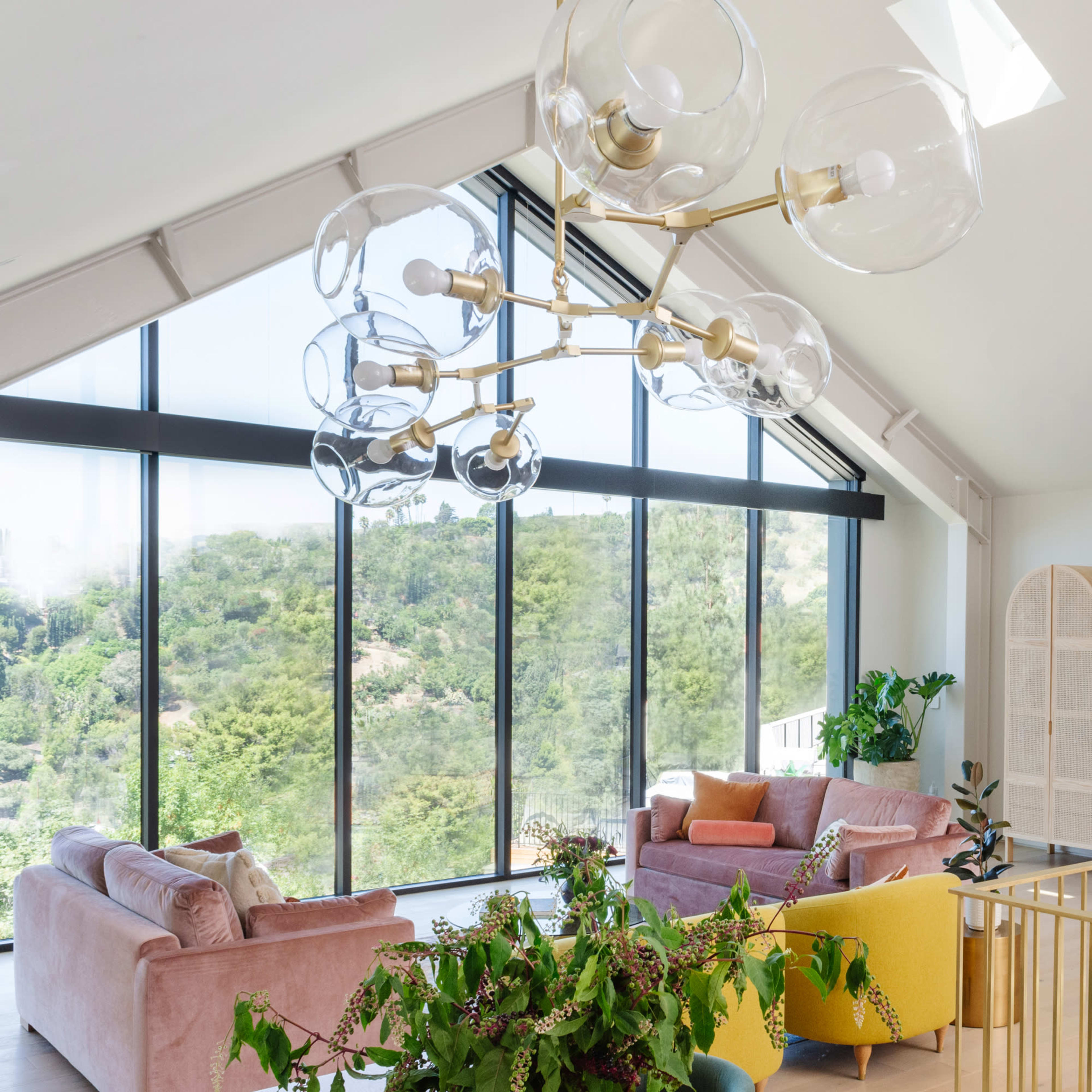 A modern living room features large windows with a view of greenery, a chandelier with glass orbs, and a mix of pink and yellow furniture.