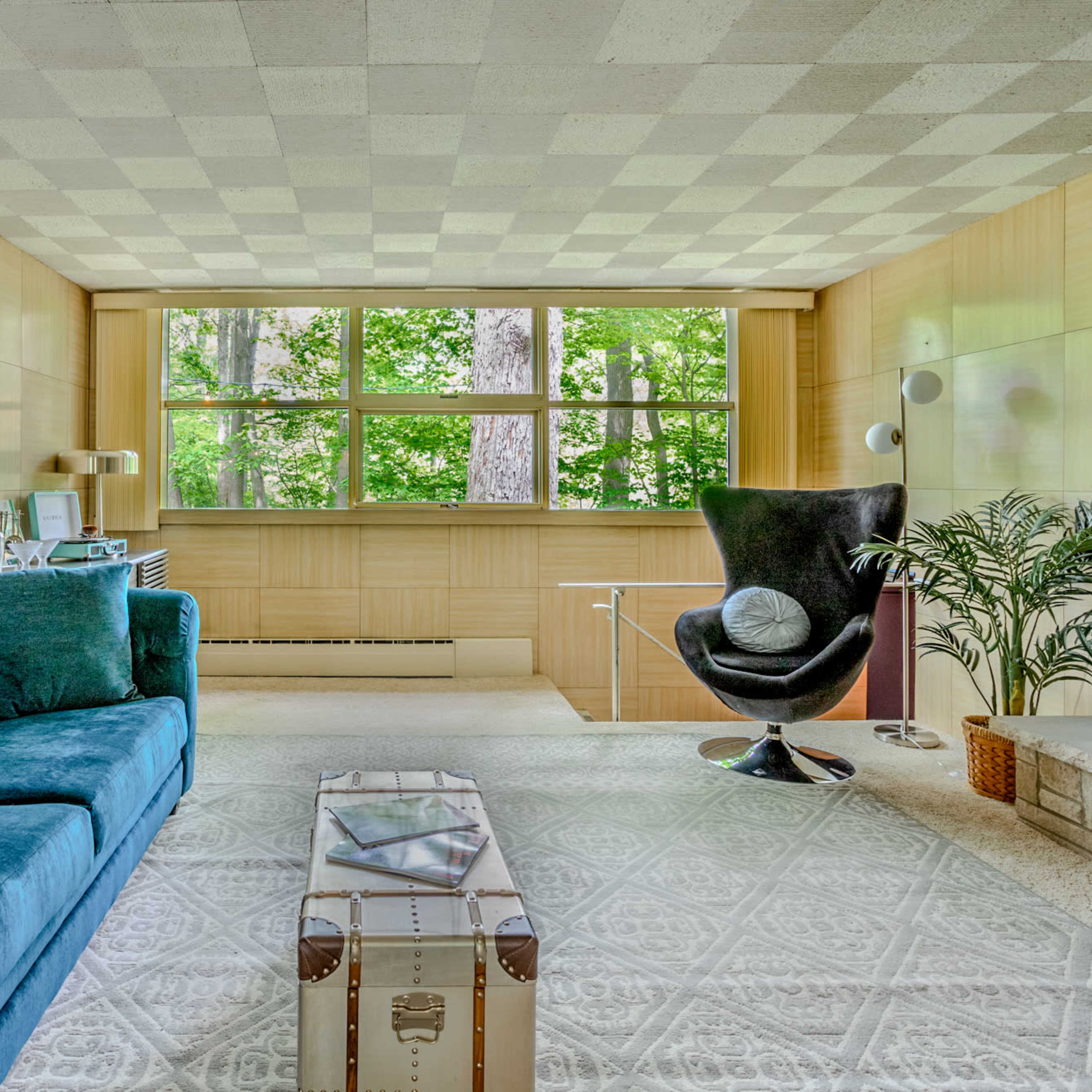 A modern interior living space featuring a blue tufted sofa, a black chair, a glass coffee table, and large windows with views of greenery outside.