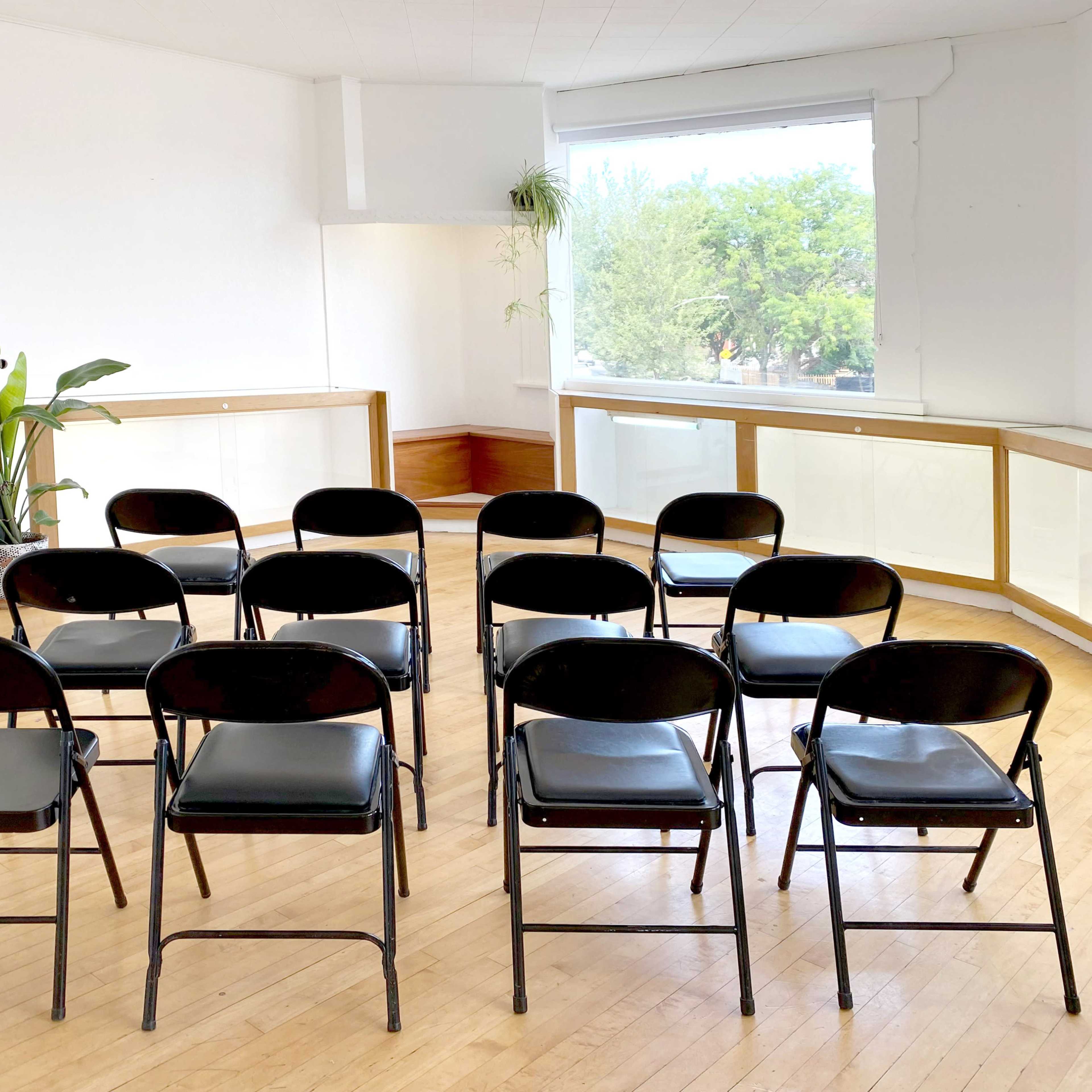 A group of black folding chairs is arranged in two rows facing a large window in a bright, empty room.