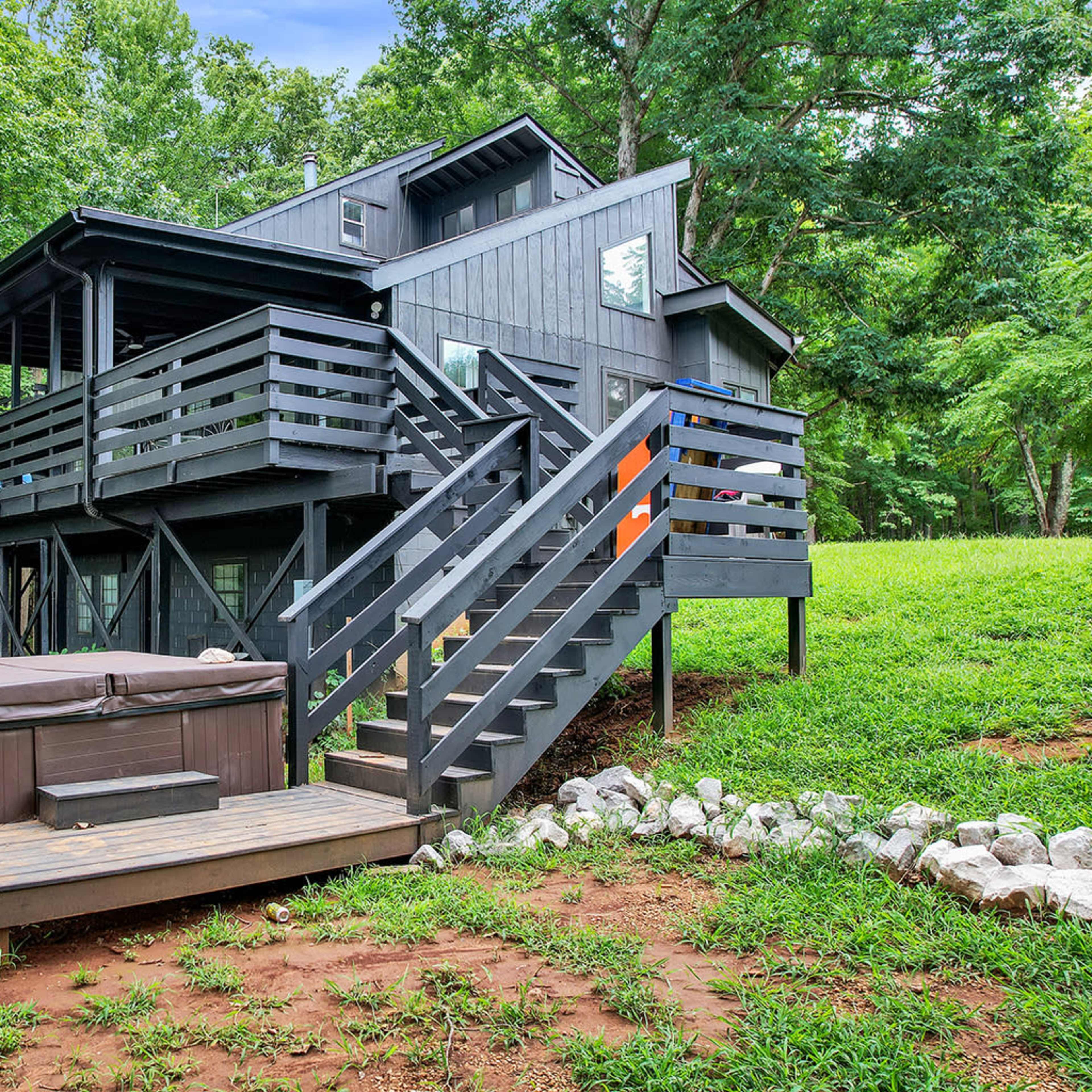 A two-story black house with a deck and hot tub is surrounded by greenery and trees.
