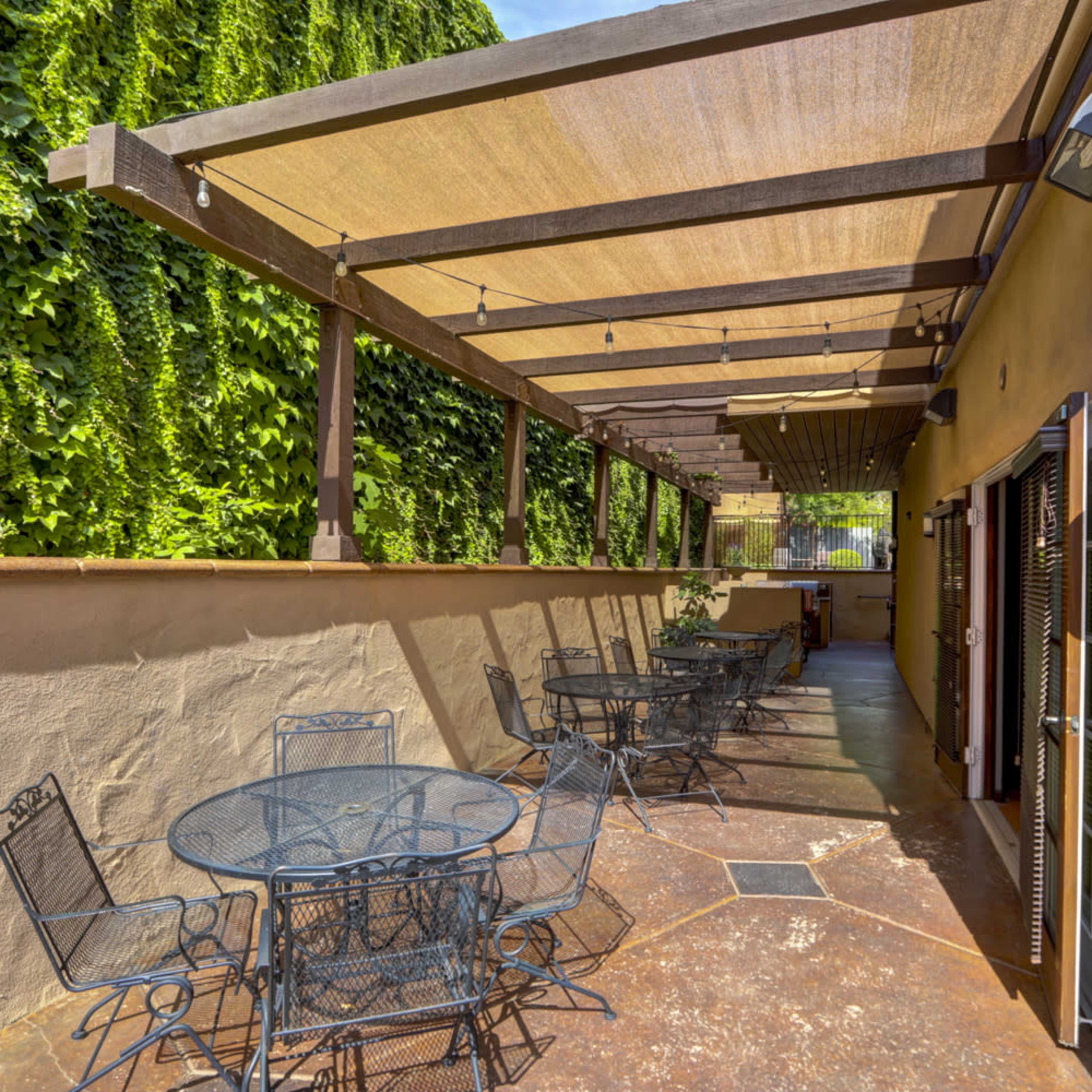 A patio area with several round tables and chairs shaded by a large canopy and surrounded by green foliage.