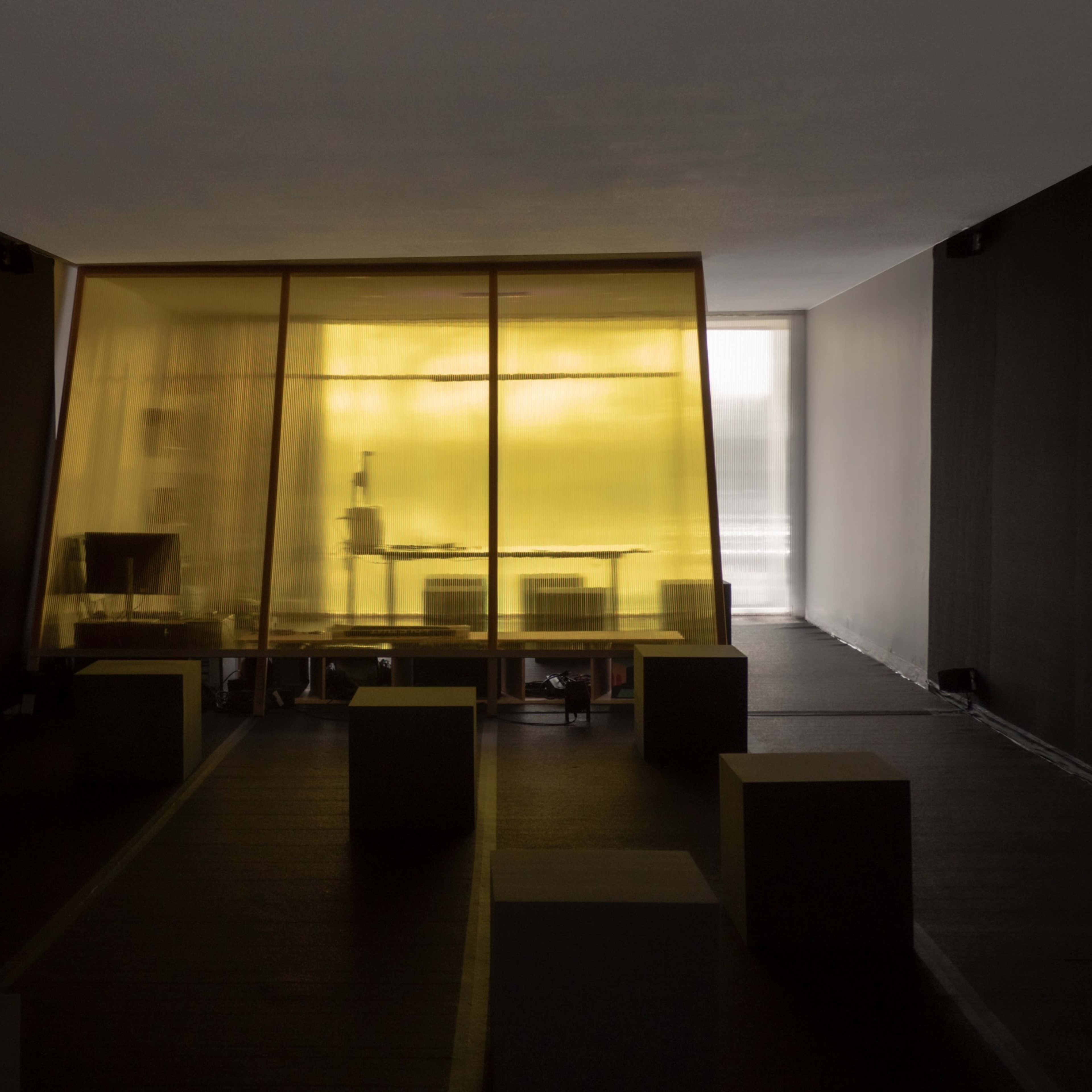 A room features a yellow-tinted glass enclosure containing a workstation, surrounded by gray, cube-shaped seating.