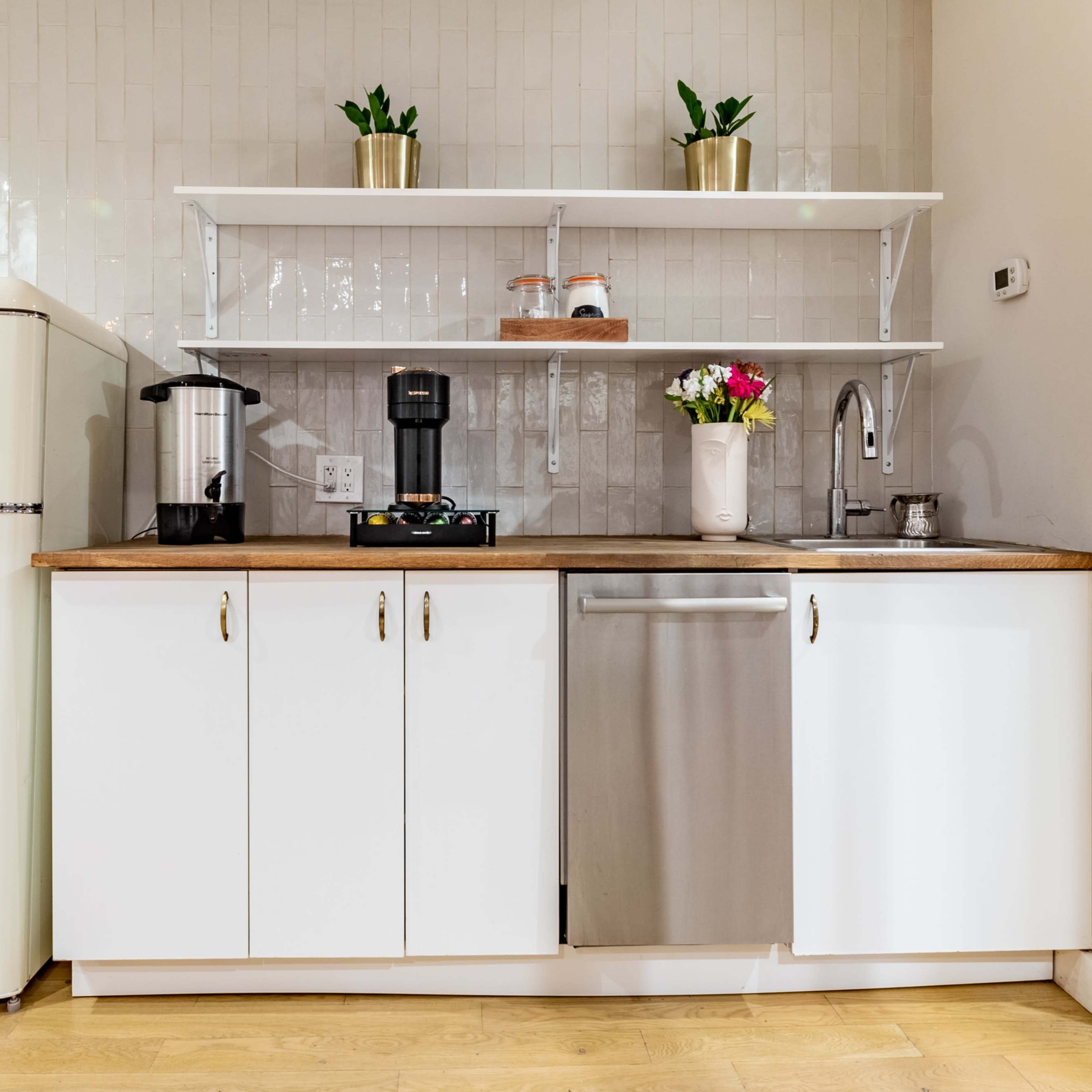 A kitchen features a retro-style refrigerator, sleek cabinetry, a coffee maker, and a stainless steel dishwasher, with plants and flowers on the shelves.
