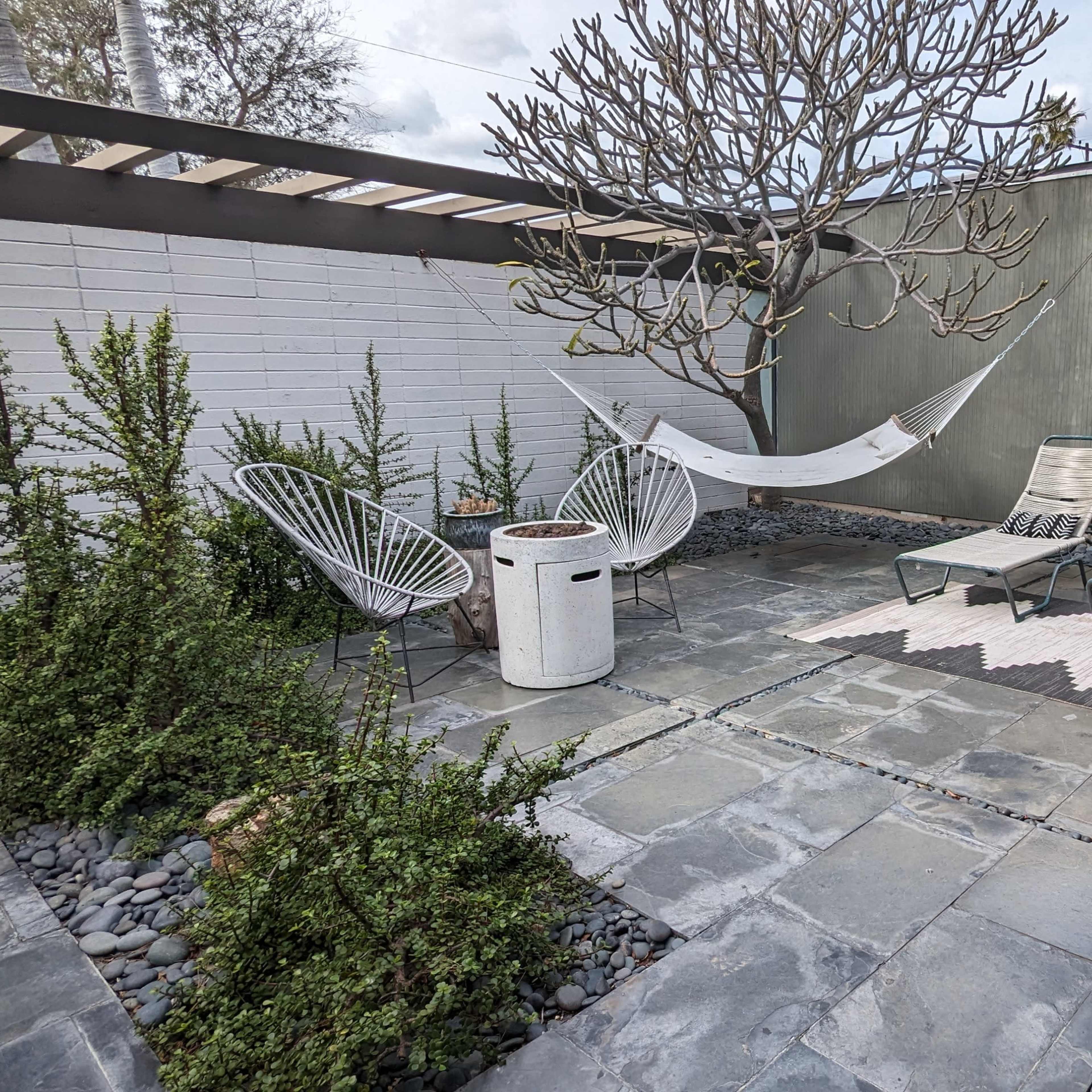 A minimalist outdoor space featuring a hammock, two lounge chairs, and a small table surrounded by stone pathways and greenery.