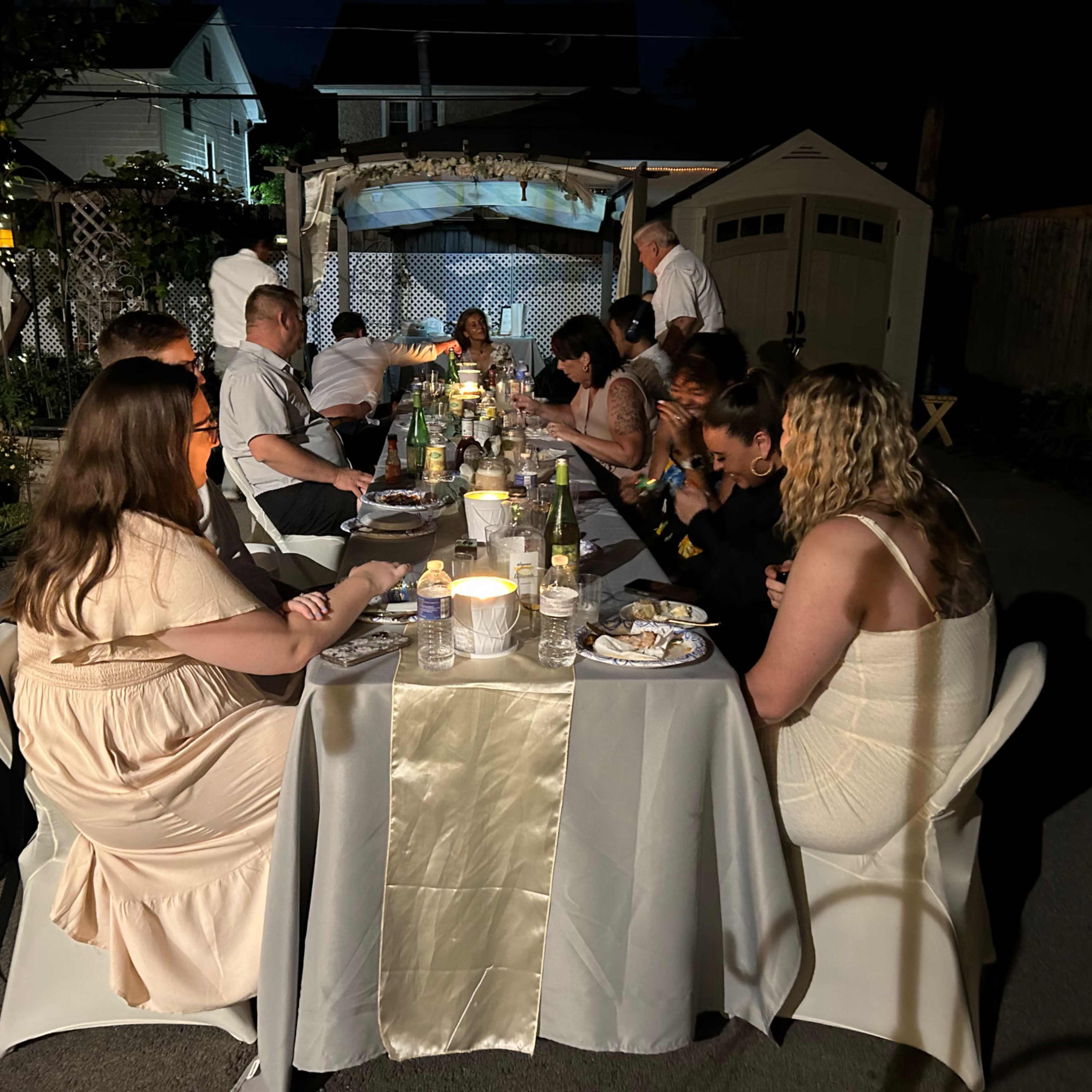 A group of people is seated at a long table in a backyard, enjoying a dinner gathering under soft lighting at night.