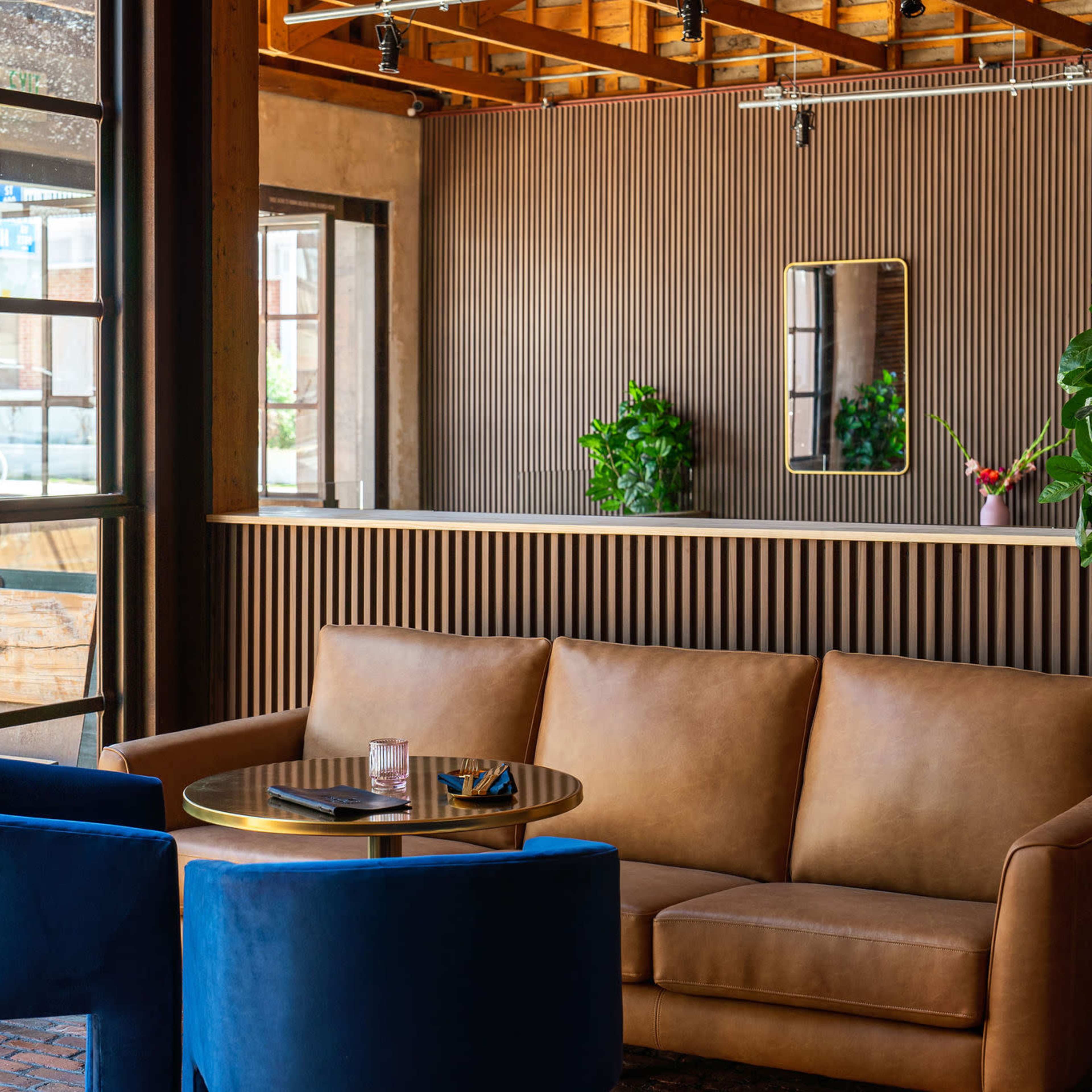 A cozy lounge area with a brown leather sofa, two blue chairs, a round table, and a wall featuring a mirror and potted plants.