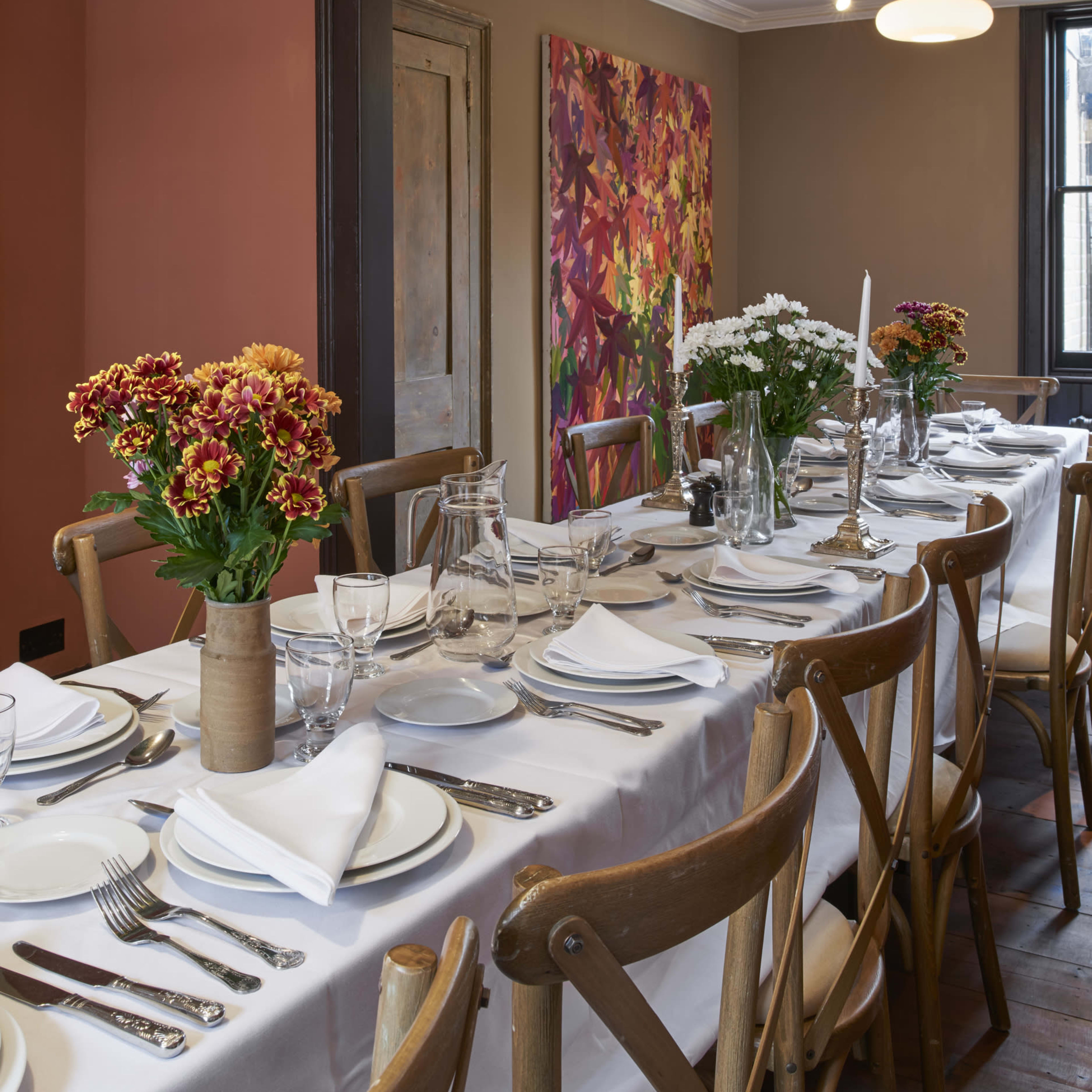 A long dining table set with white tablecloth, ceramic dishes, glassware, and adorned with bouquets of flowers is positioned in a room featuring colorful artwork on the wall.