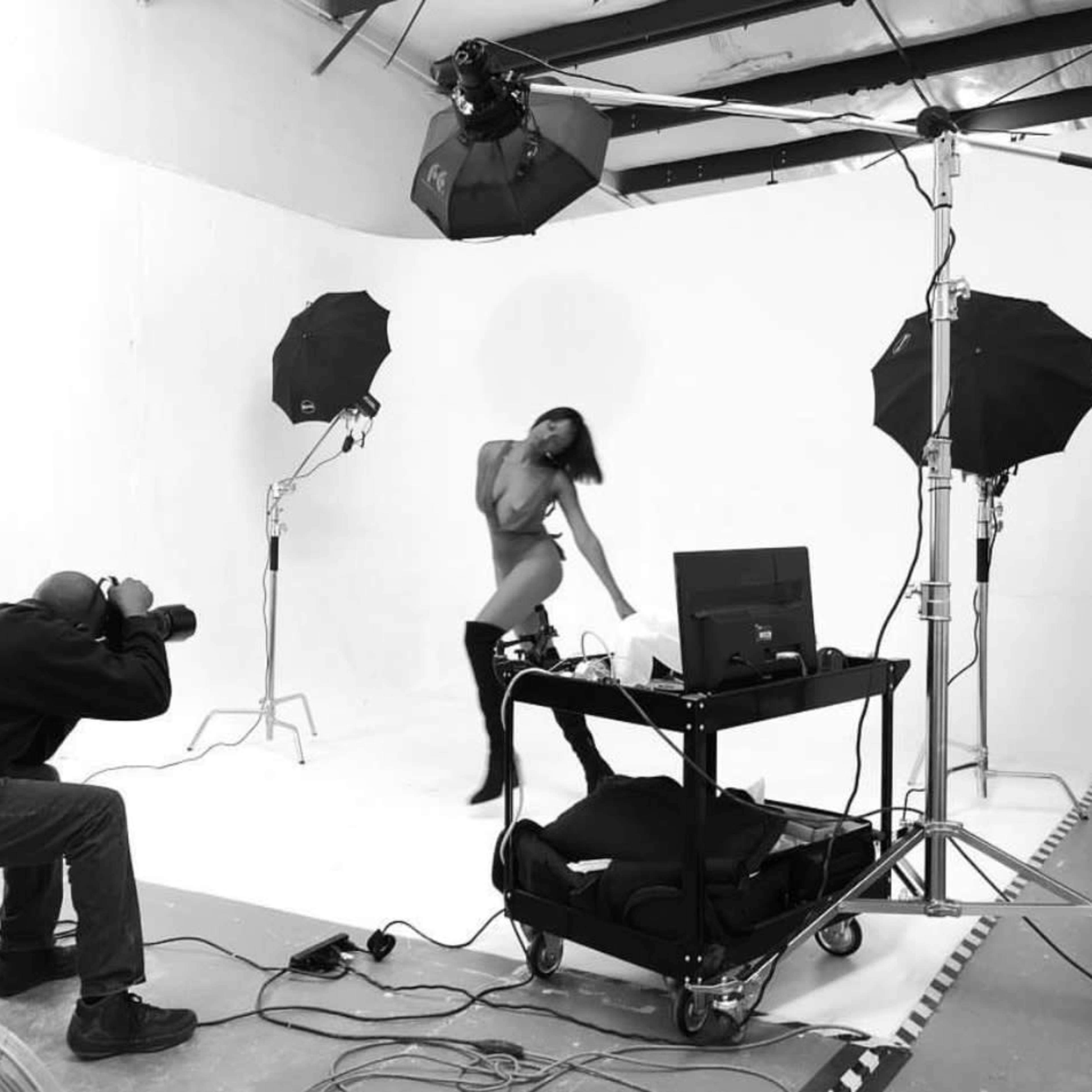 A photographer captures a model posing in a studio with white backdrops and lighting equipment.