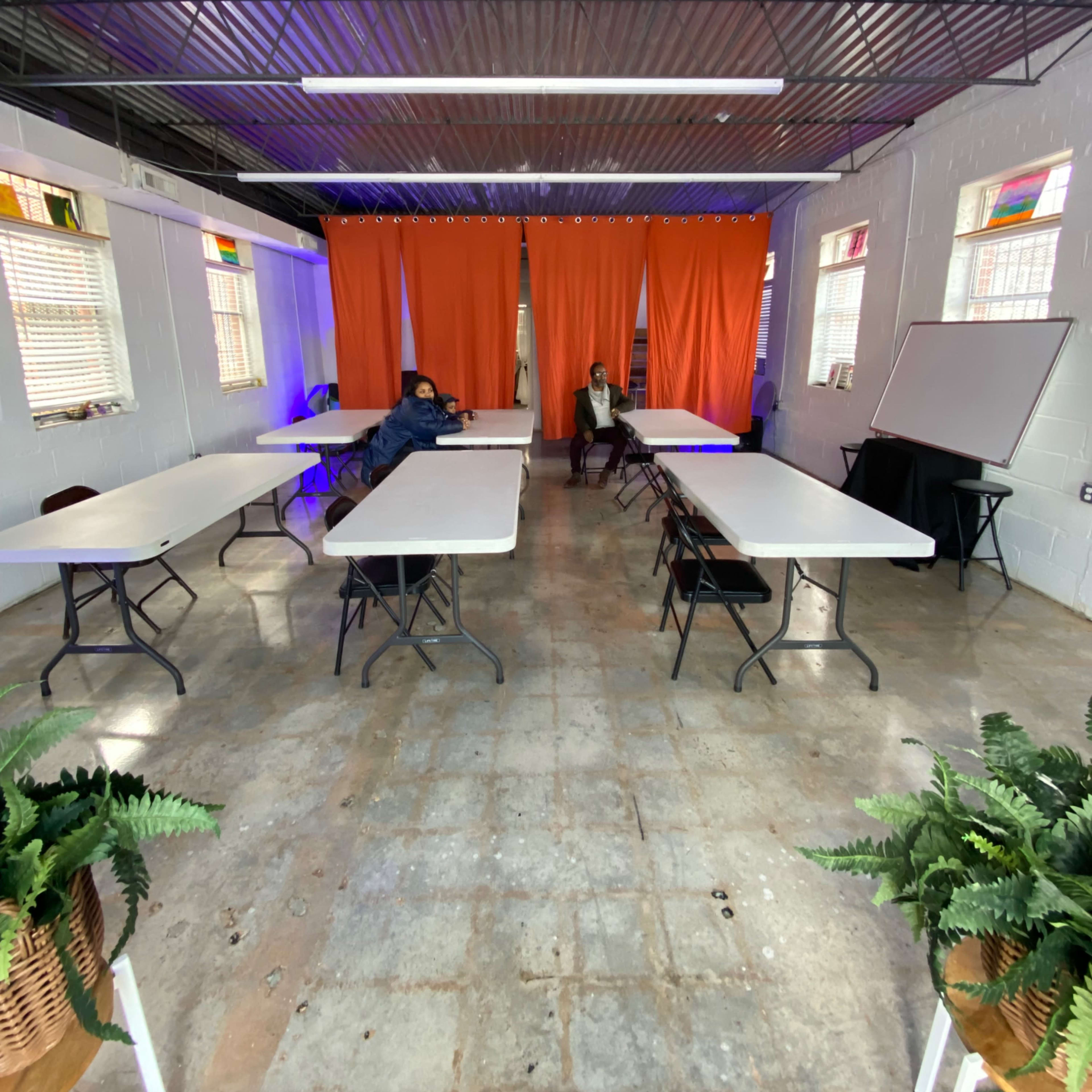A sparsely furnished room with several white tables arranged in rows, two people seated at the tables, and orange curtains separating the space.