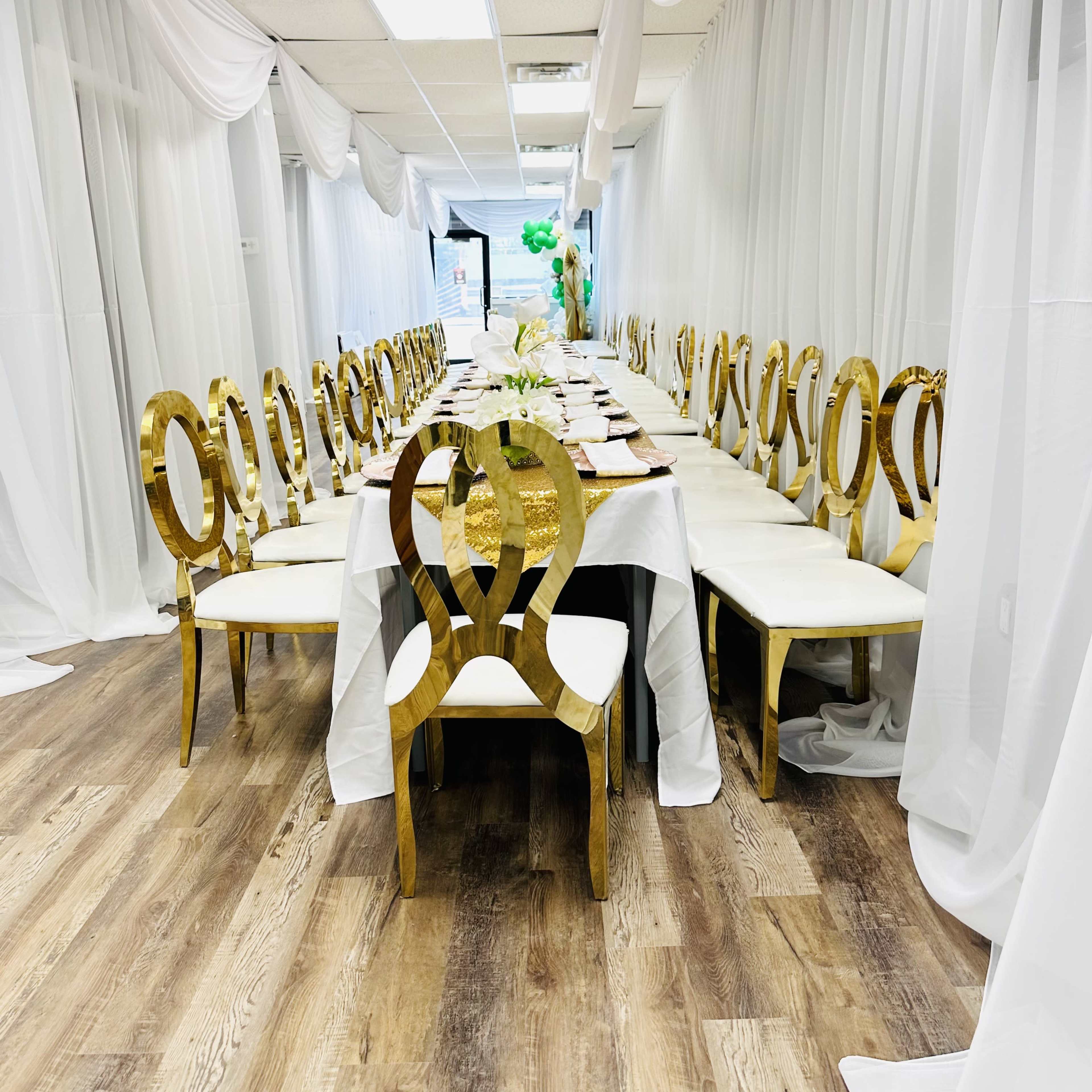 A long dining table is set with white tableware and surrounded by gold chairs, all under draped white fabric in a hallway.