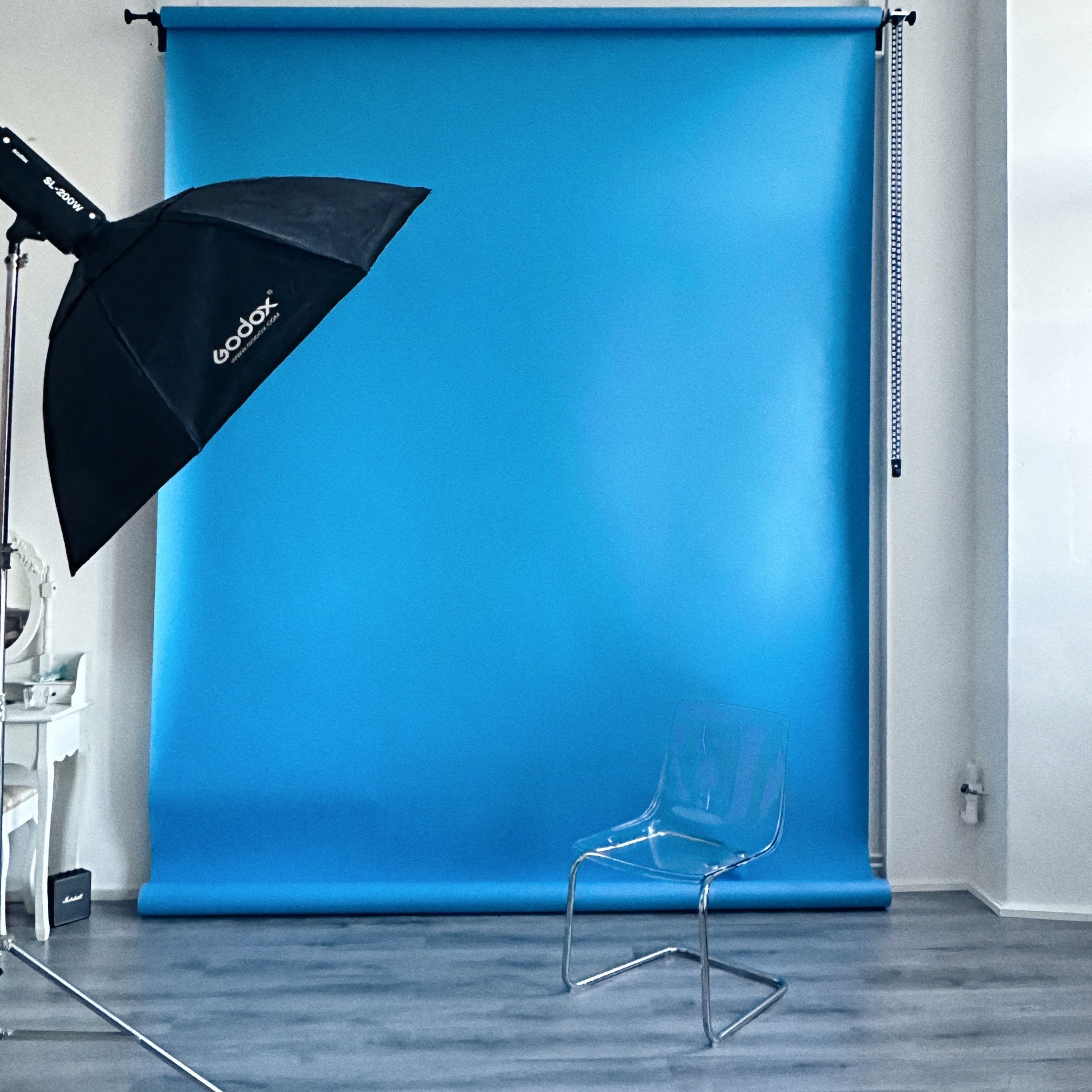 A photography studio with a blue backdrop, an umbrella softbox light, and a transparent chair on a wooden floor.