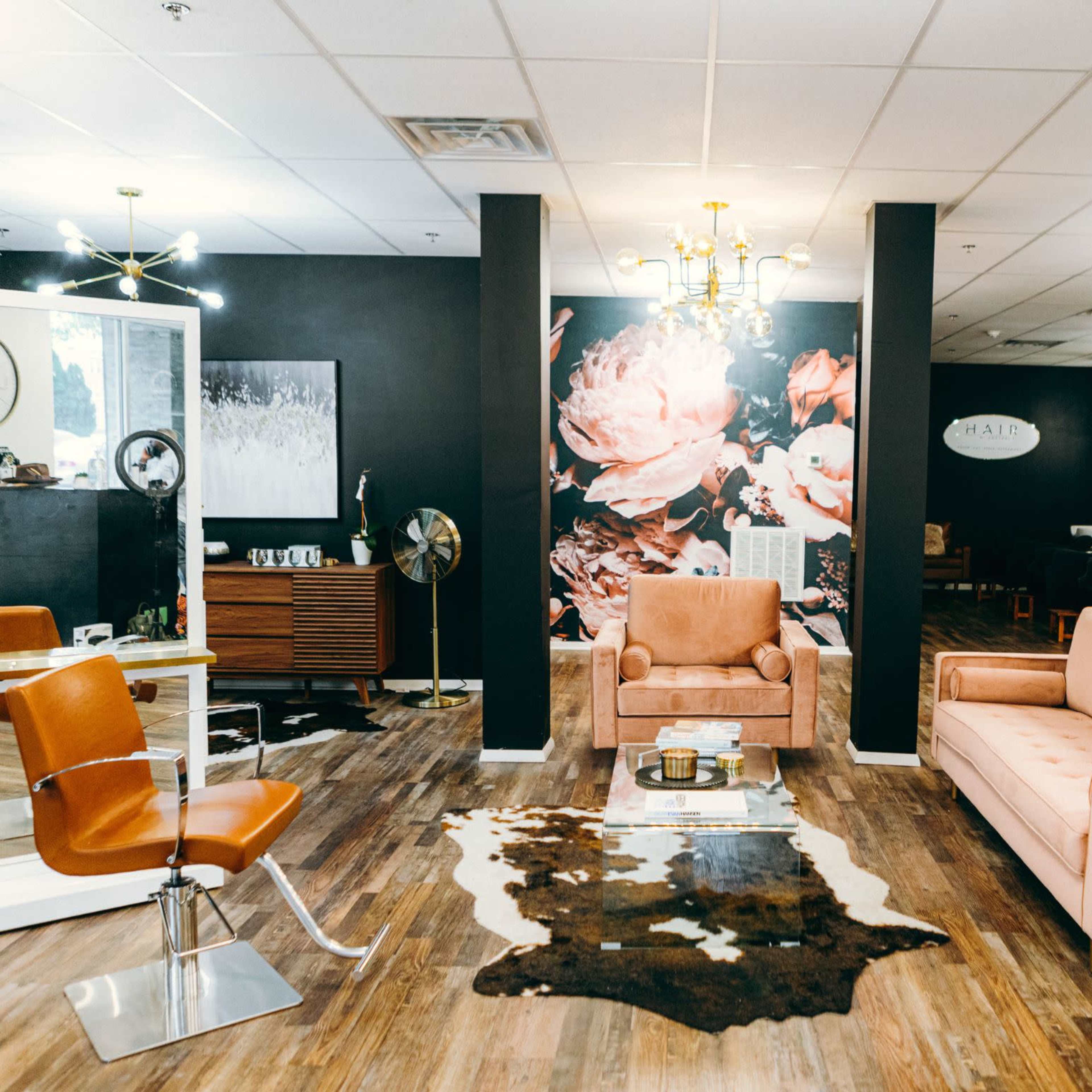 A modern salon features a pink velvet sofa, a cowhide rug, and a wall adorned with floral artwork, set against dark walls and bright lighting.
