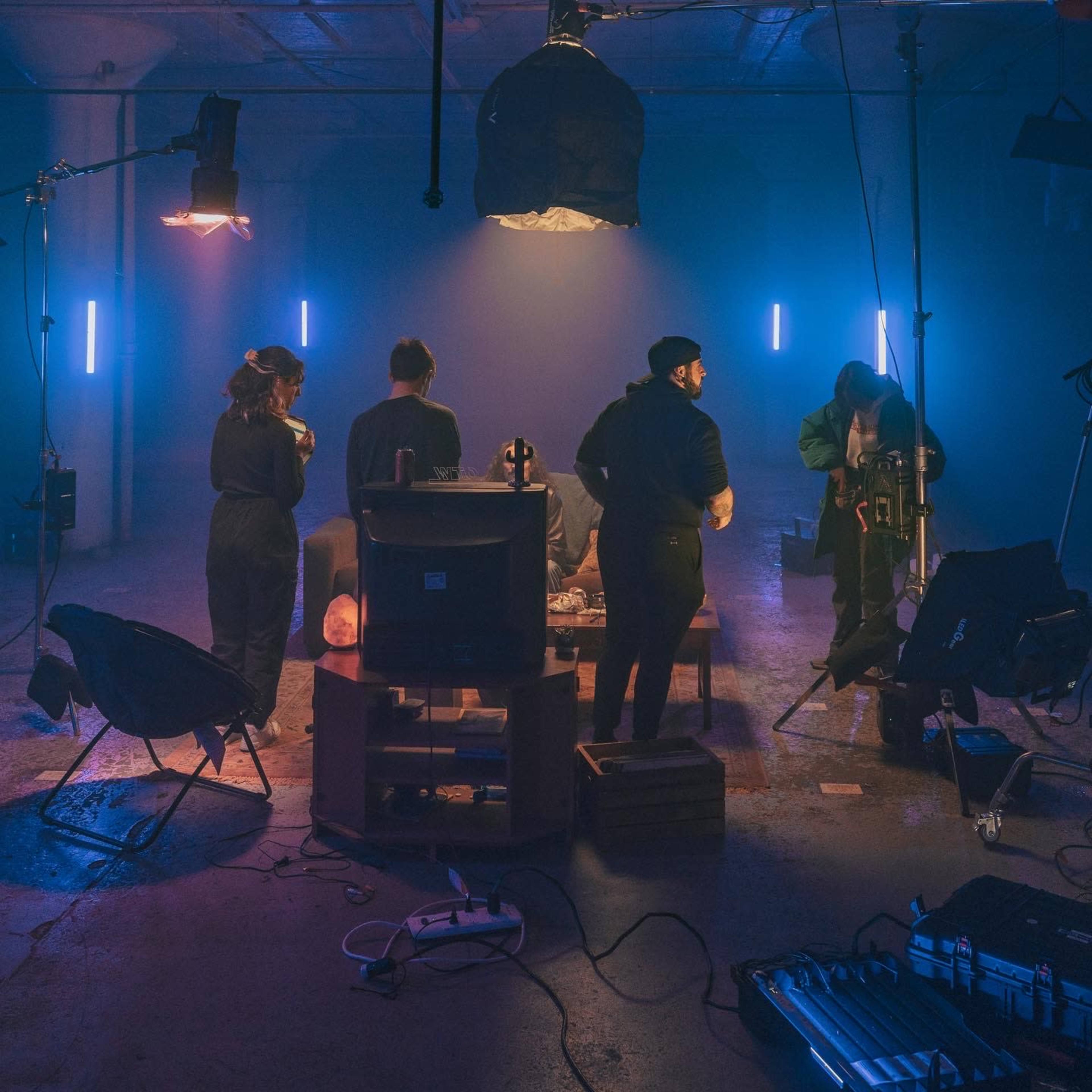 A group of five individuals in dark clothing is arranged around a low table in a dimly lit room with blue and purple lighting, setting up equipment for a project.