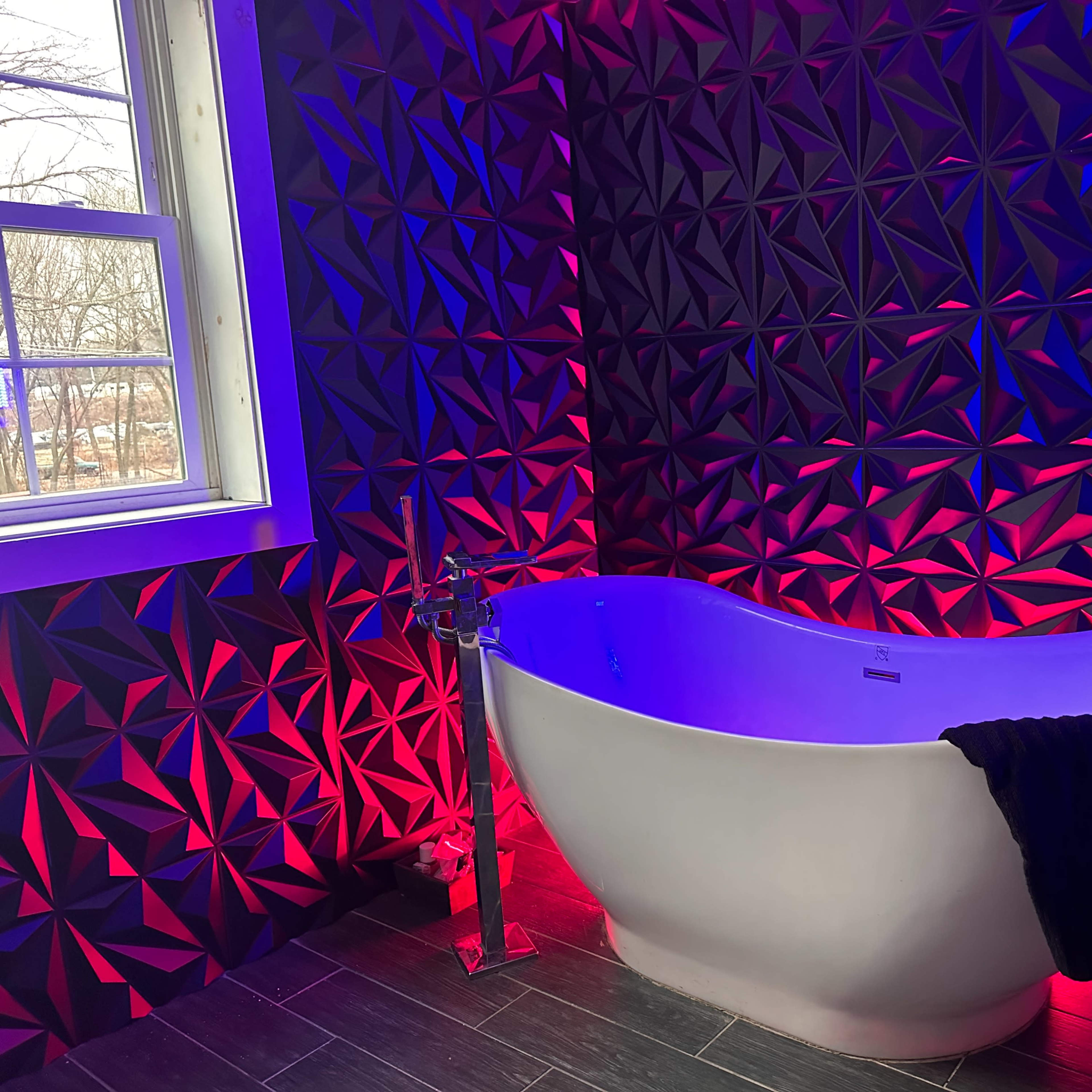 A freestanding white bathtub is positioned against a textured wall illuminated with red and blue lights, with a window nearby.