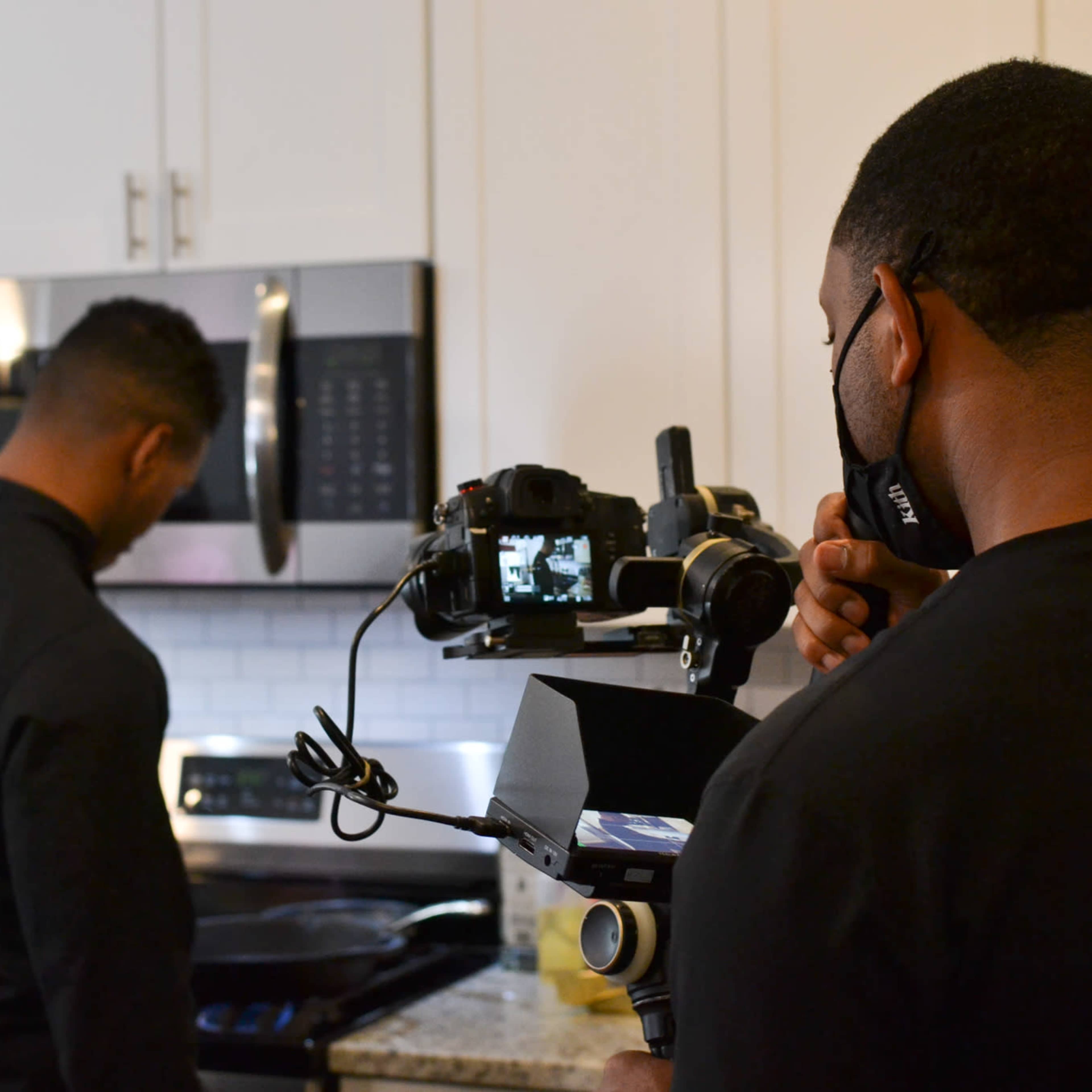 A person in a chef's jacket is cooking on a stovetop while another individual operates a camera and stabilizer in a kitchen setting.