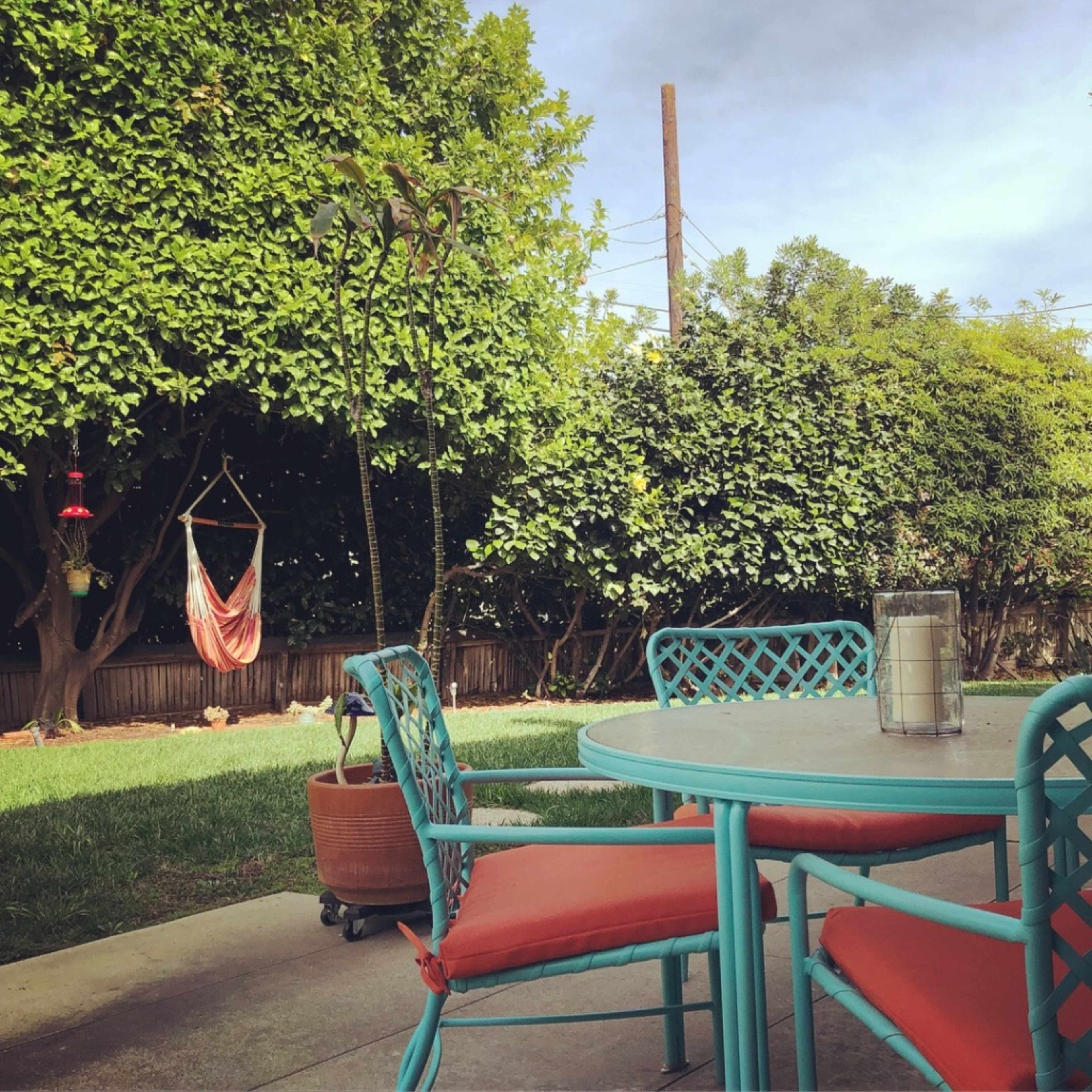 A patio scene features a round table with teal chairs and orange cushions, surrounded by lush greenery and a hanging hammock in the background.