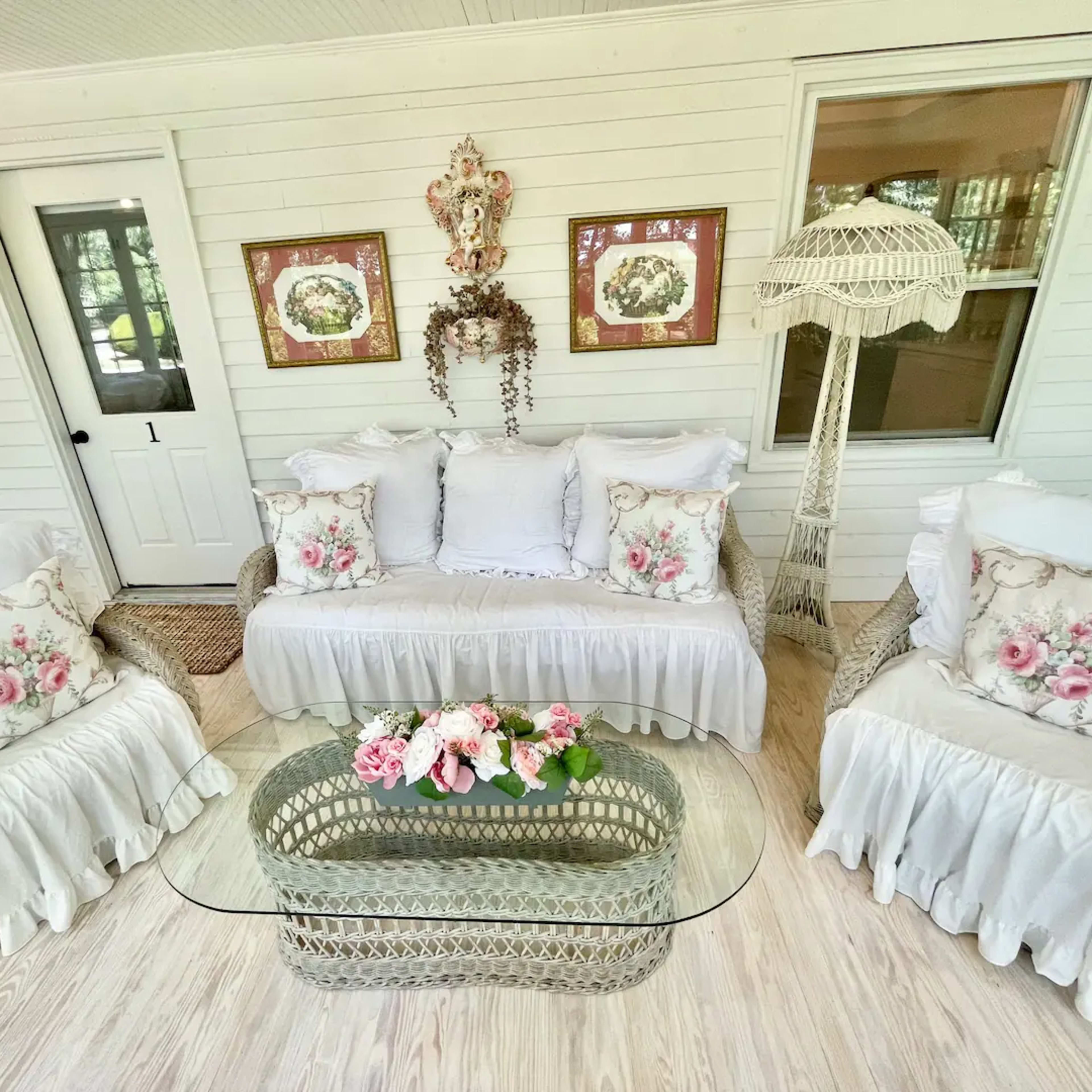 A light-filled sunroom featuring a rattan sofa with white floral pillows and a glass coffee table adorned with a floral arrangement.