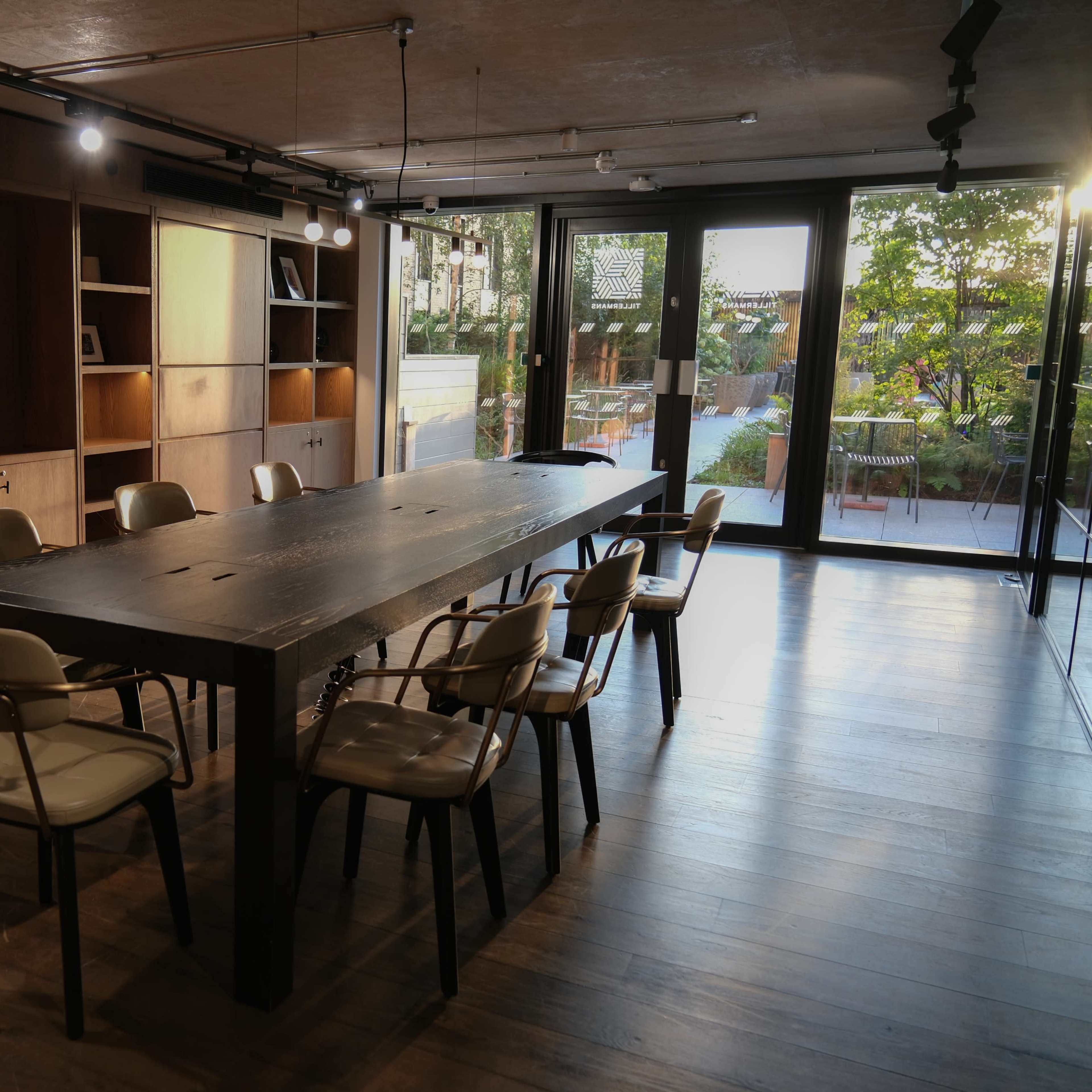 A large, wooden conference table surrounded by chairs sits in a well-lit meeting room with floor-to-ceiling windows overlooking a garden.