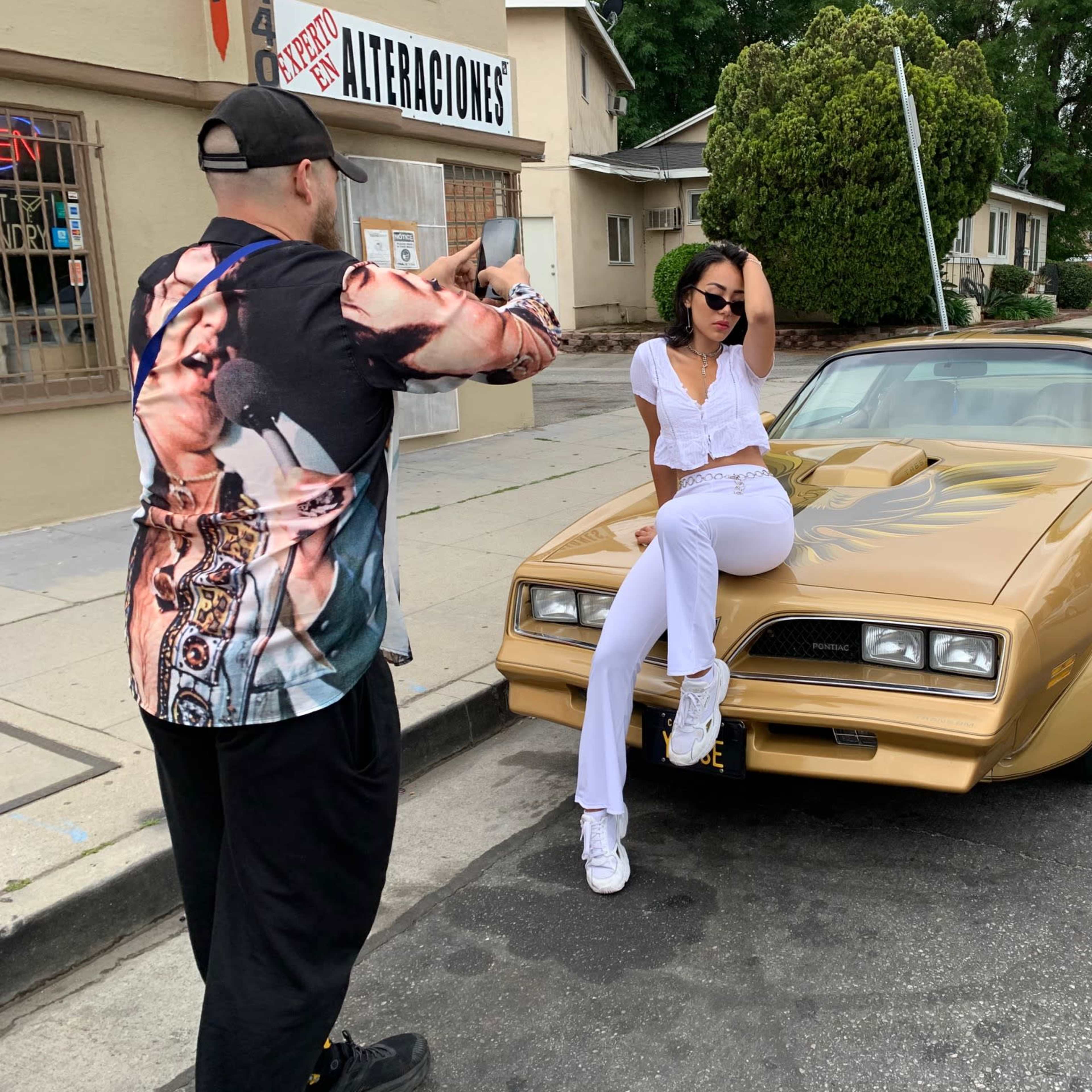 A photographer takes pictures of a model posing on a gold car on a street in front of a storefront.