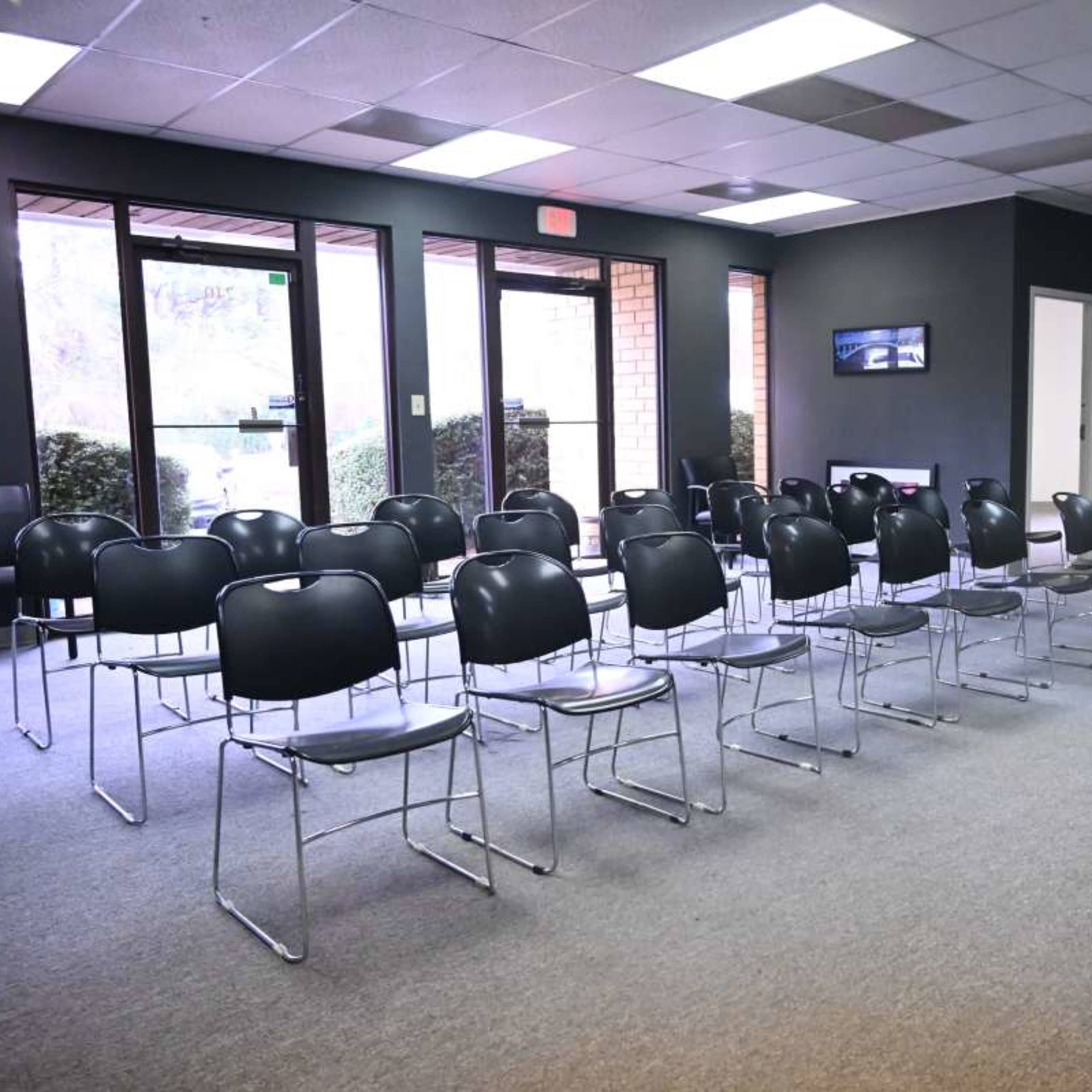 A waiting room with several rows of metal chairs arranged in front of a TV and a single armchair in the corner.