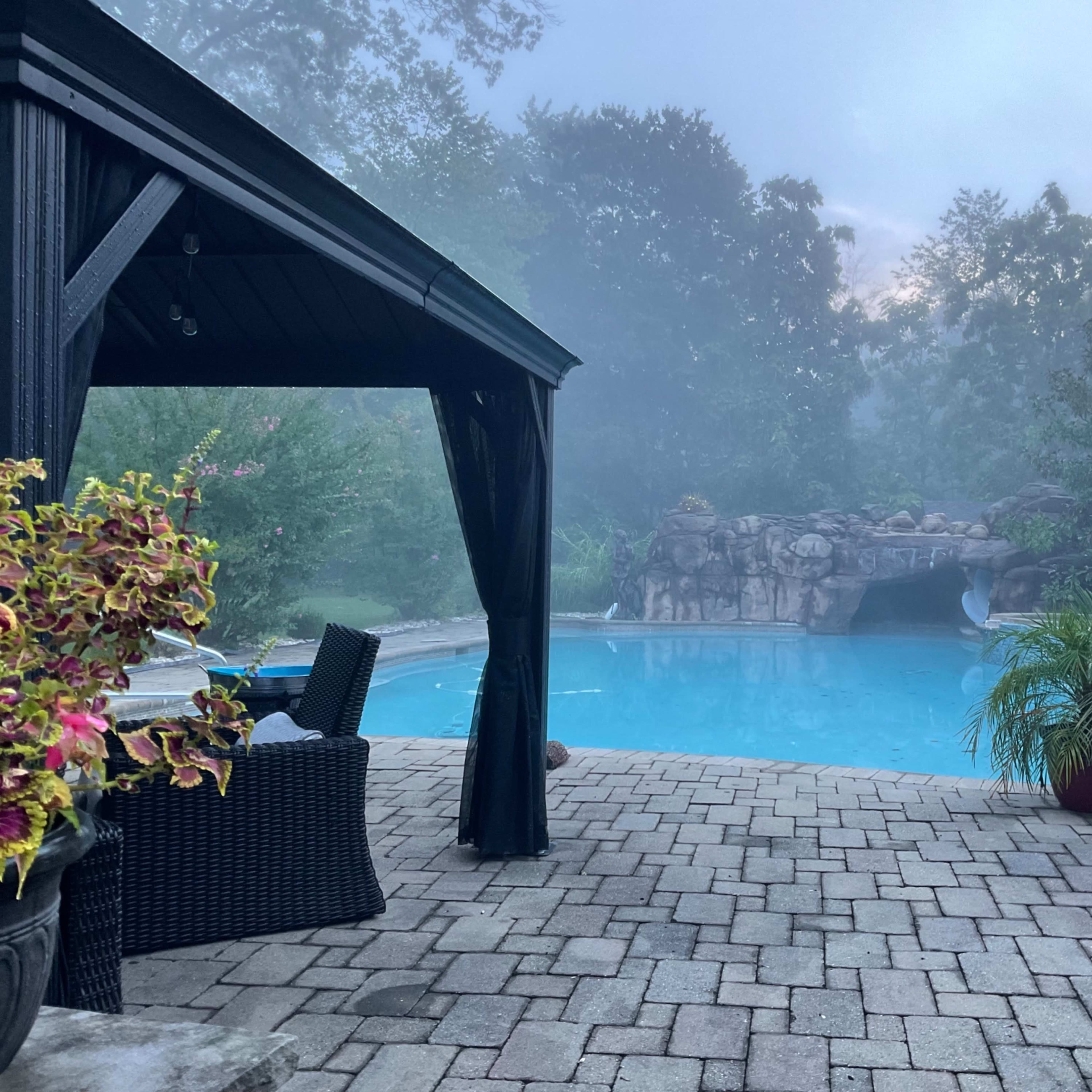 A misty morning scene features a blue swimming pool surrounded by stone pavers, with a gazebo and potted plants nearby.