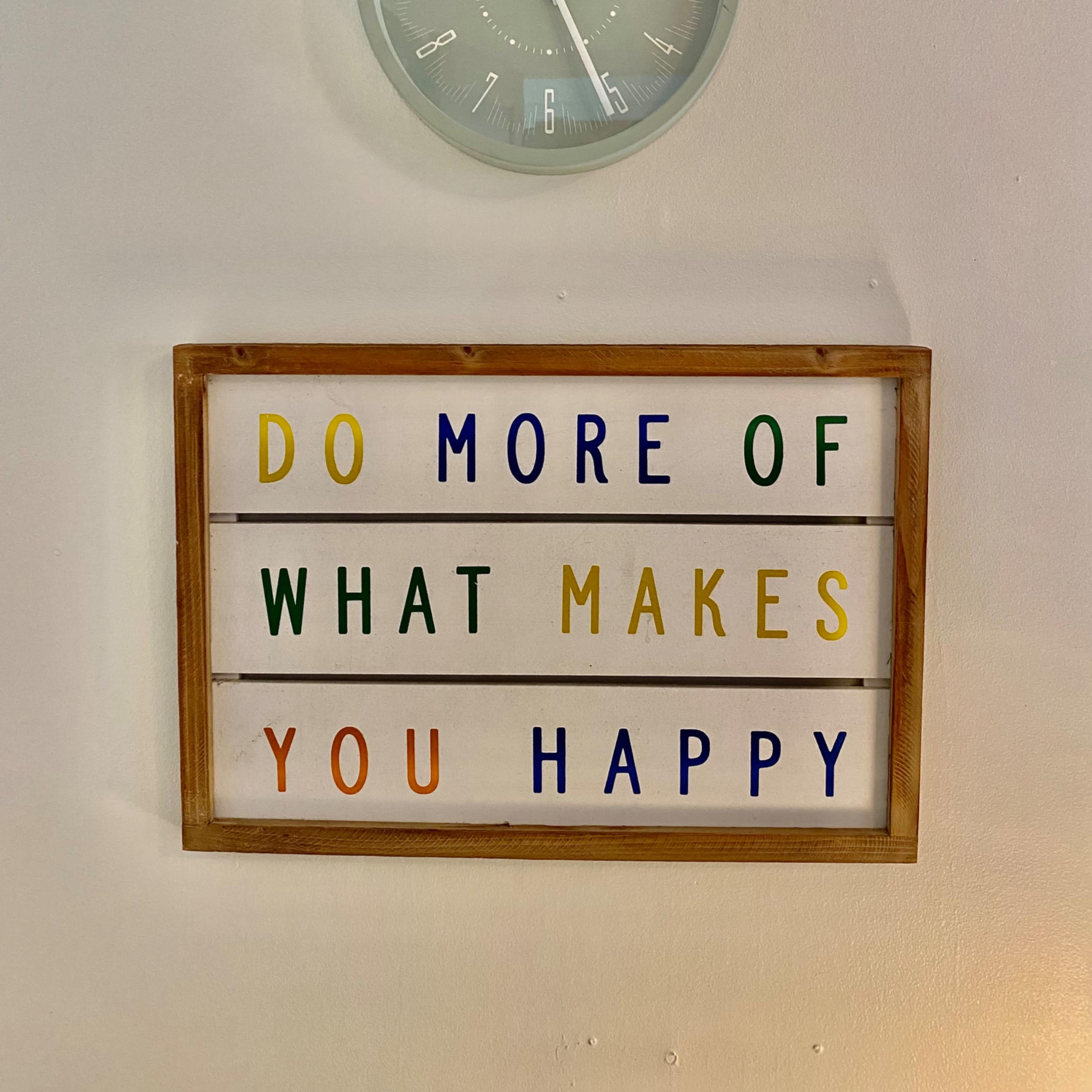 A wooden sign with colorful text that reads, "DO MORE OF WHAT MAKES YOU HAPPY," positioned beneath a round wall clock.